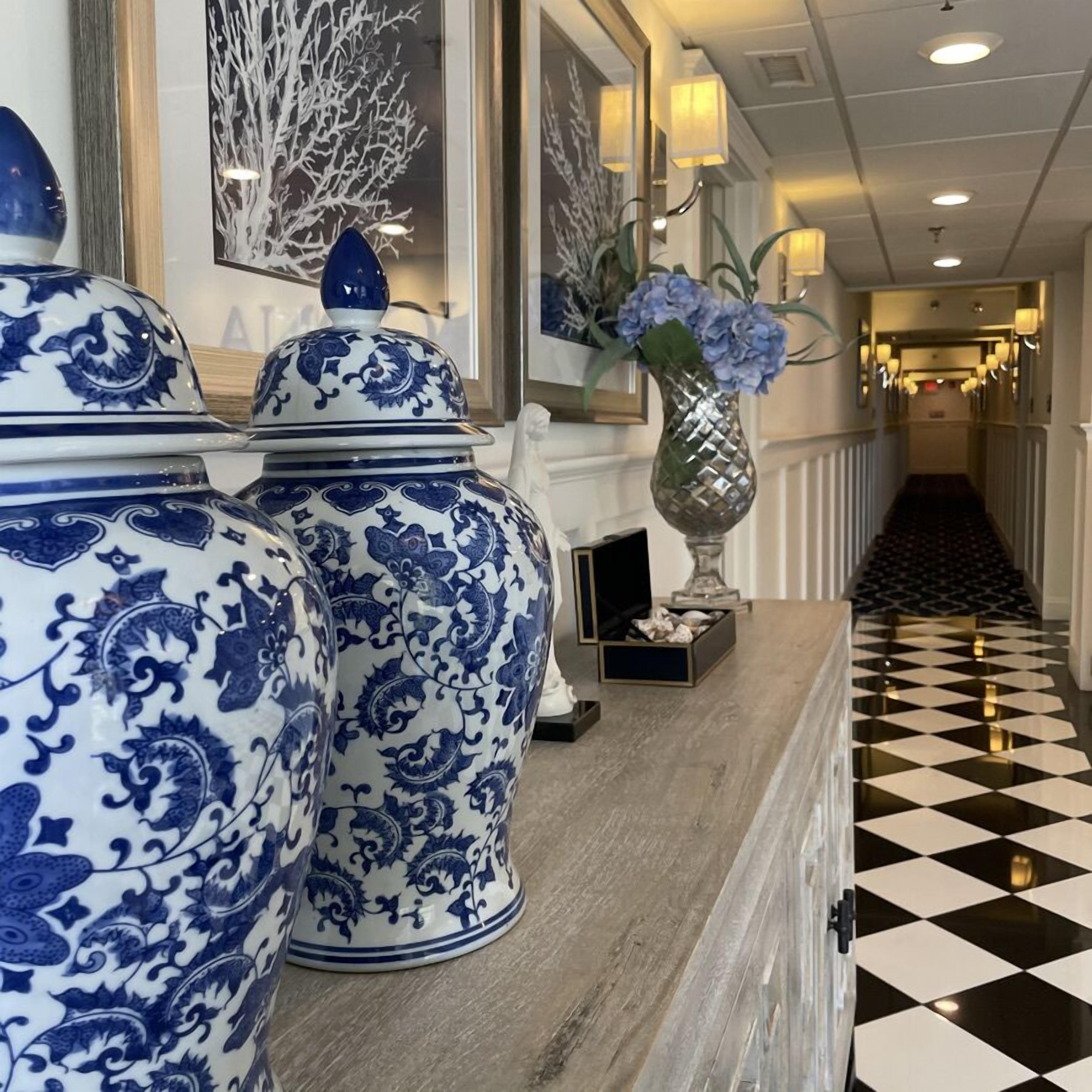Un pasillo con piso a cuadros, un jarrón con flores azules y dos jarrones grandes con estampado azul y blanco.