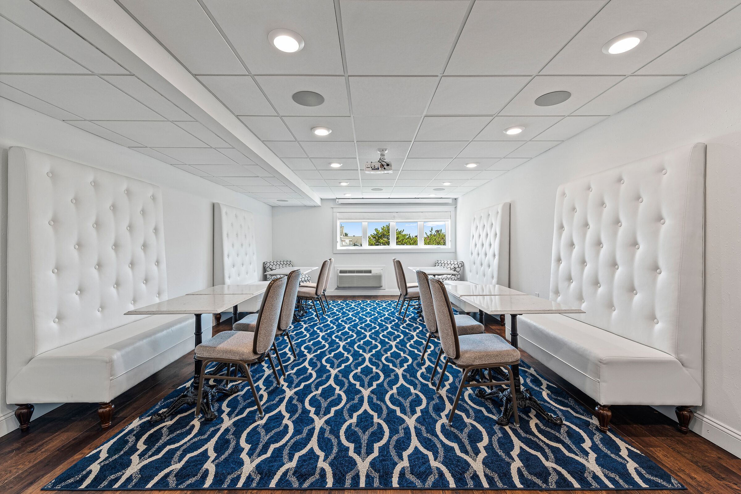 A conference room with white upholstered headboards, a blue patterned carpet, and a view of the outdoors through large windows.