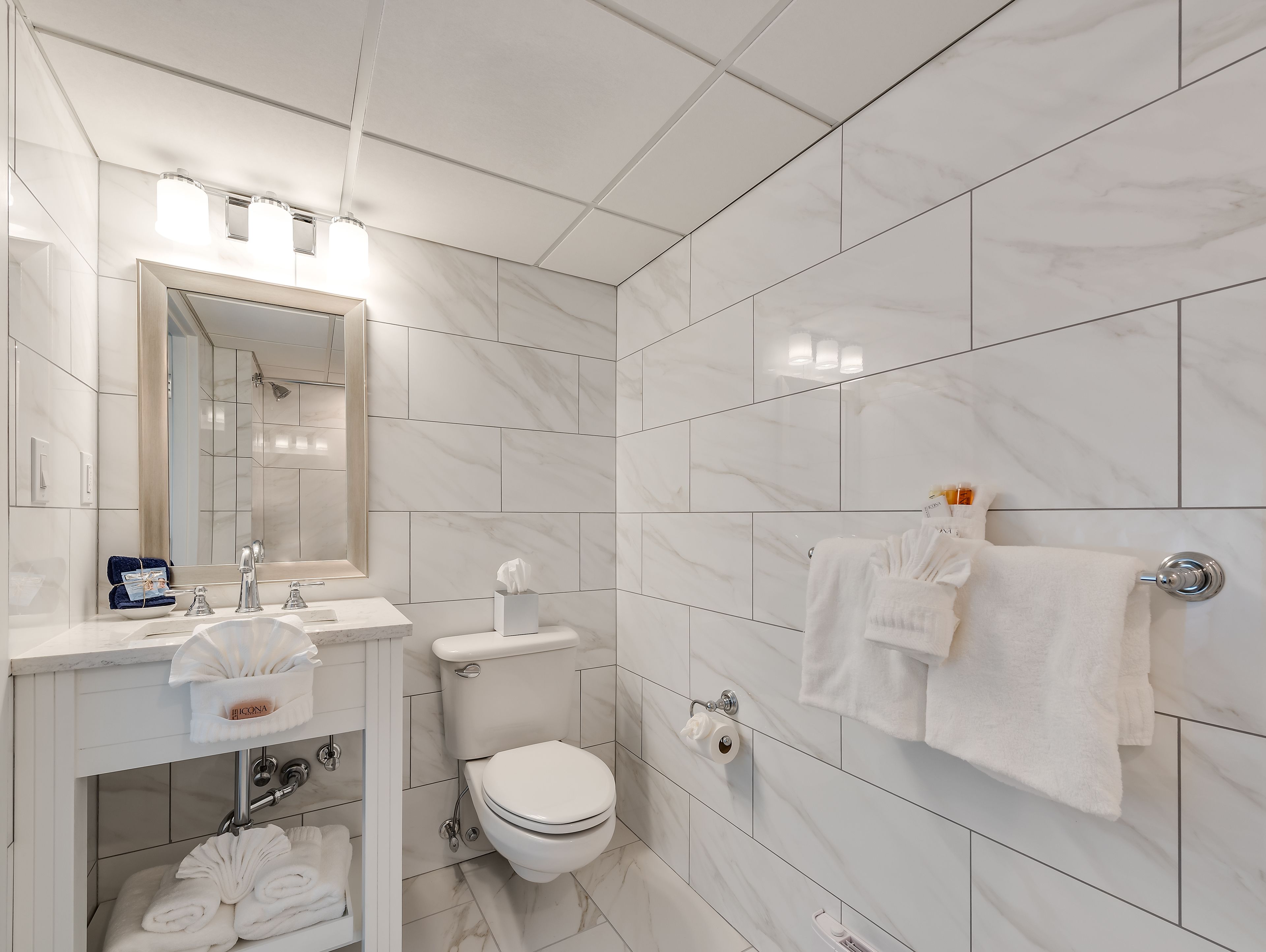 A modern bathroom with a white marble tile design, a large mirror, a sink with a towel holder, and a toilet.