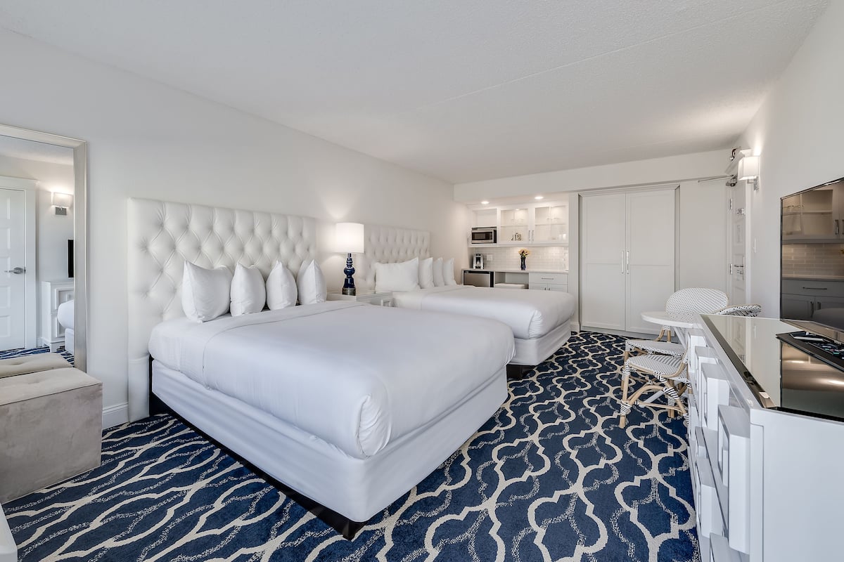 A hotel room with a large bed, a smaller bed, a desk, a chair, a vanity, and a kitchenette.