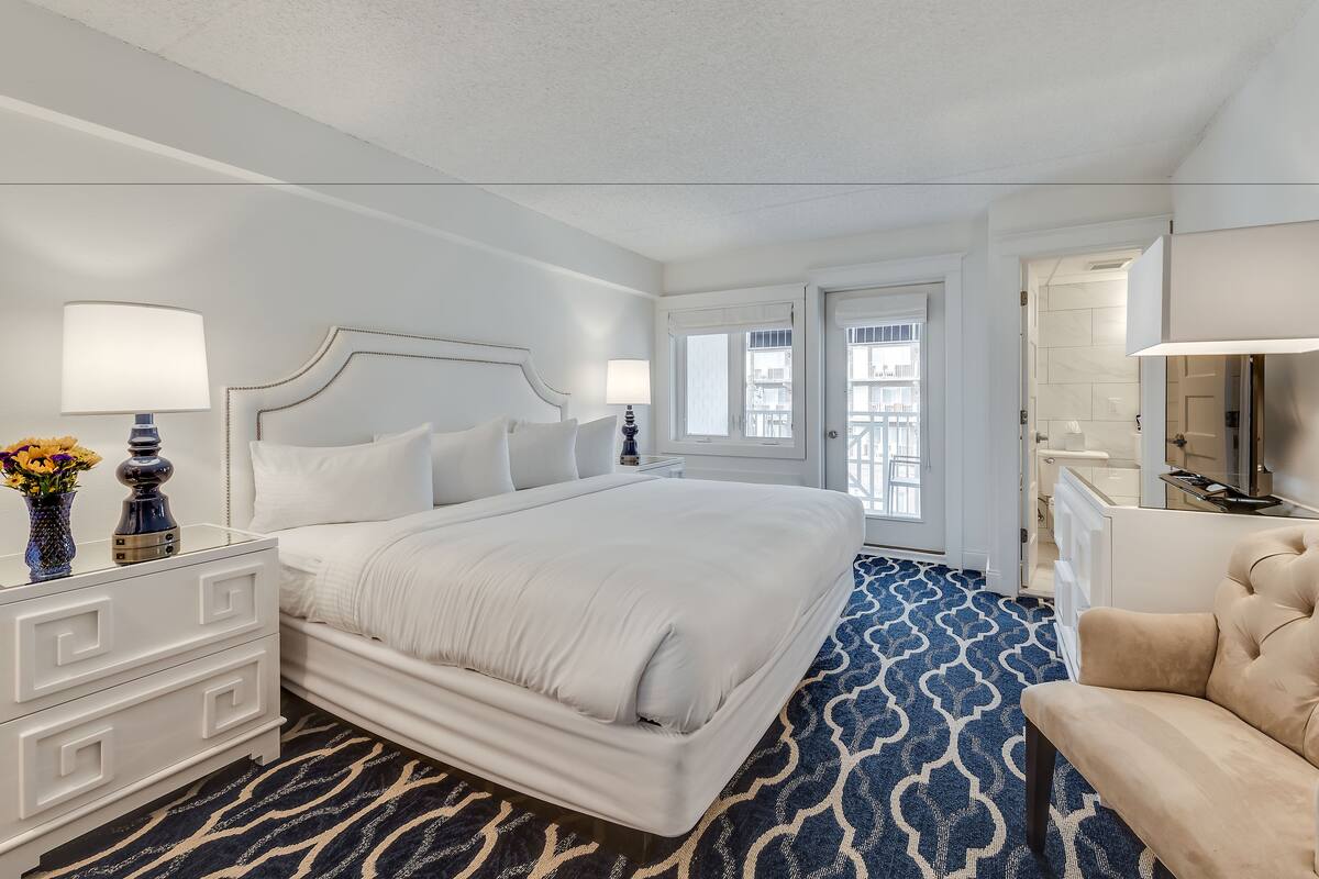 A hotel room with a large bed, a bedside table with a lamp, a chair, a bathroom with a mirror, and a view of the city through the window.