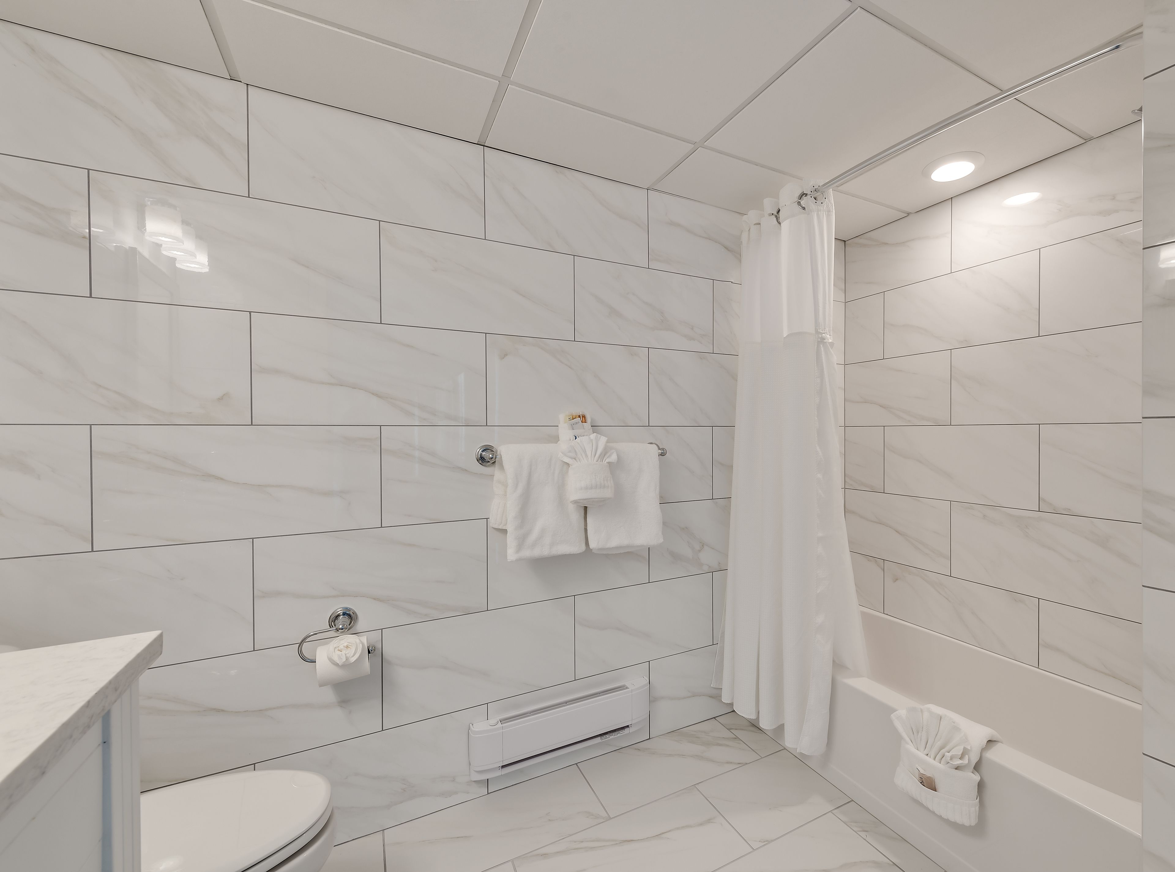 A modern bathroom with white marble tiles, a glass shower enclosure, and a toilet with a paper towel dispenser.