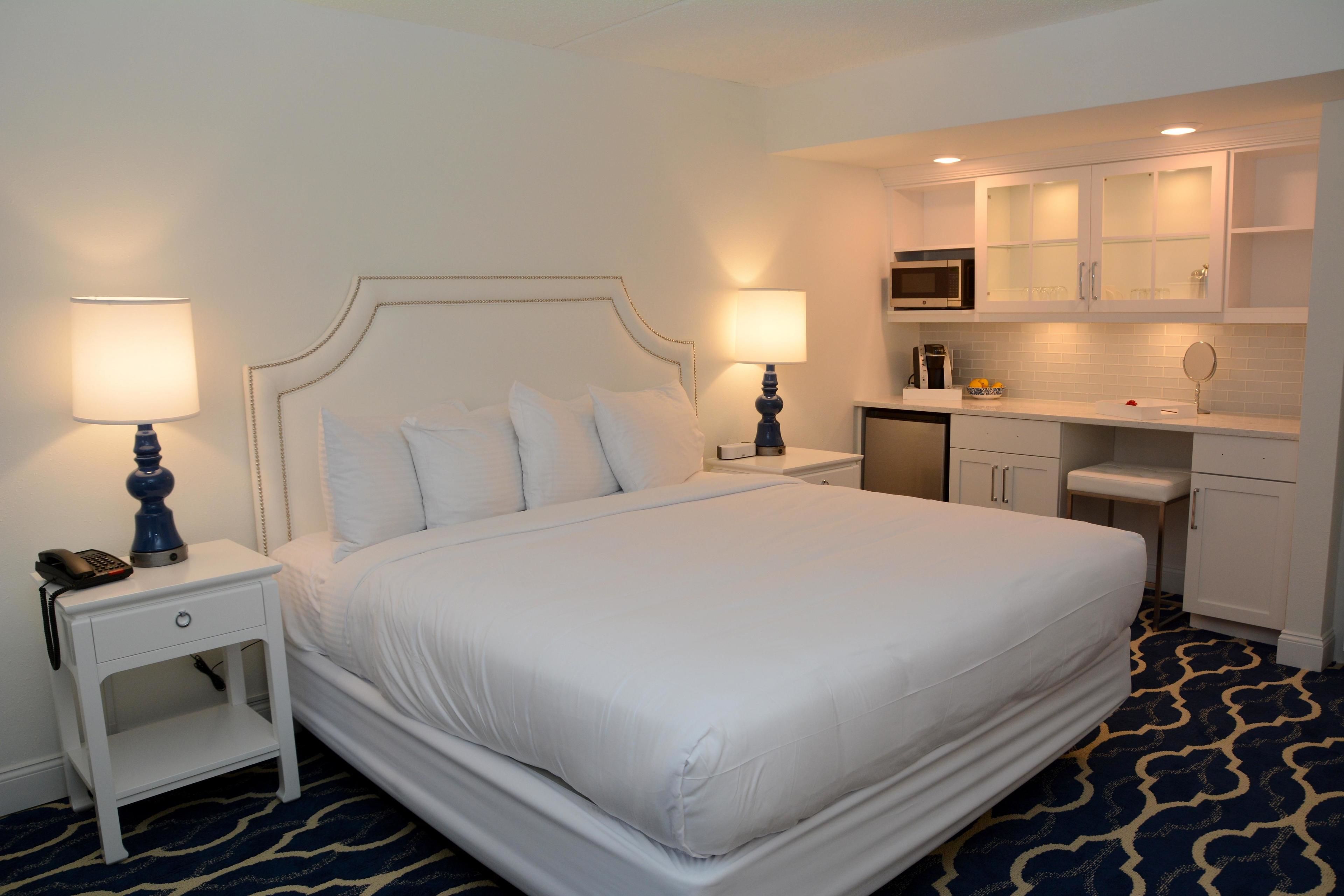 A hotel room with a large bed, two bedside tables with lamps, a small kitchenette, and a bathroom.