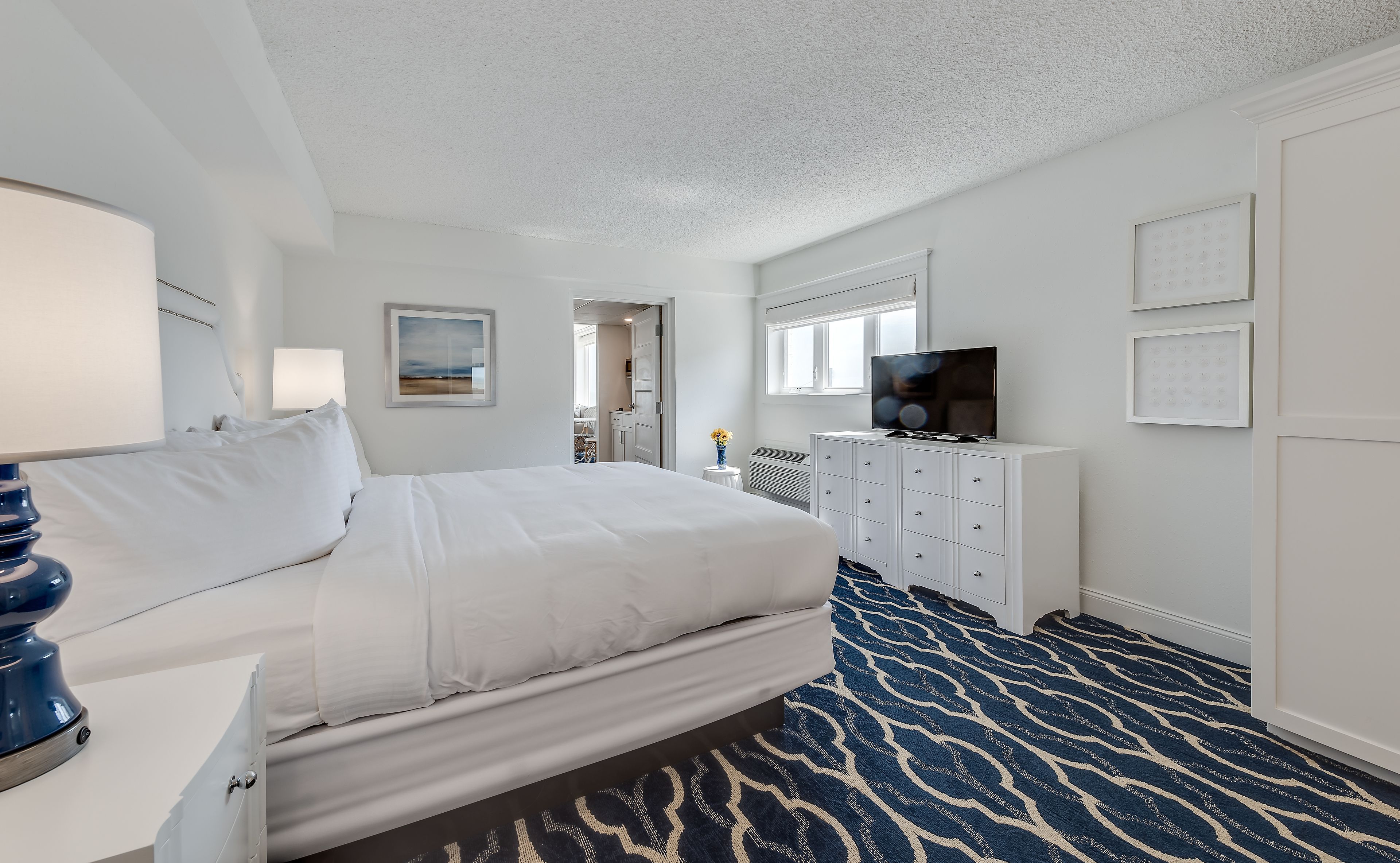 A hotel room with a large bed, a TV on a white cabinet, a patterned carpet, and a framed picture on the wall.