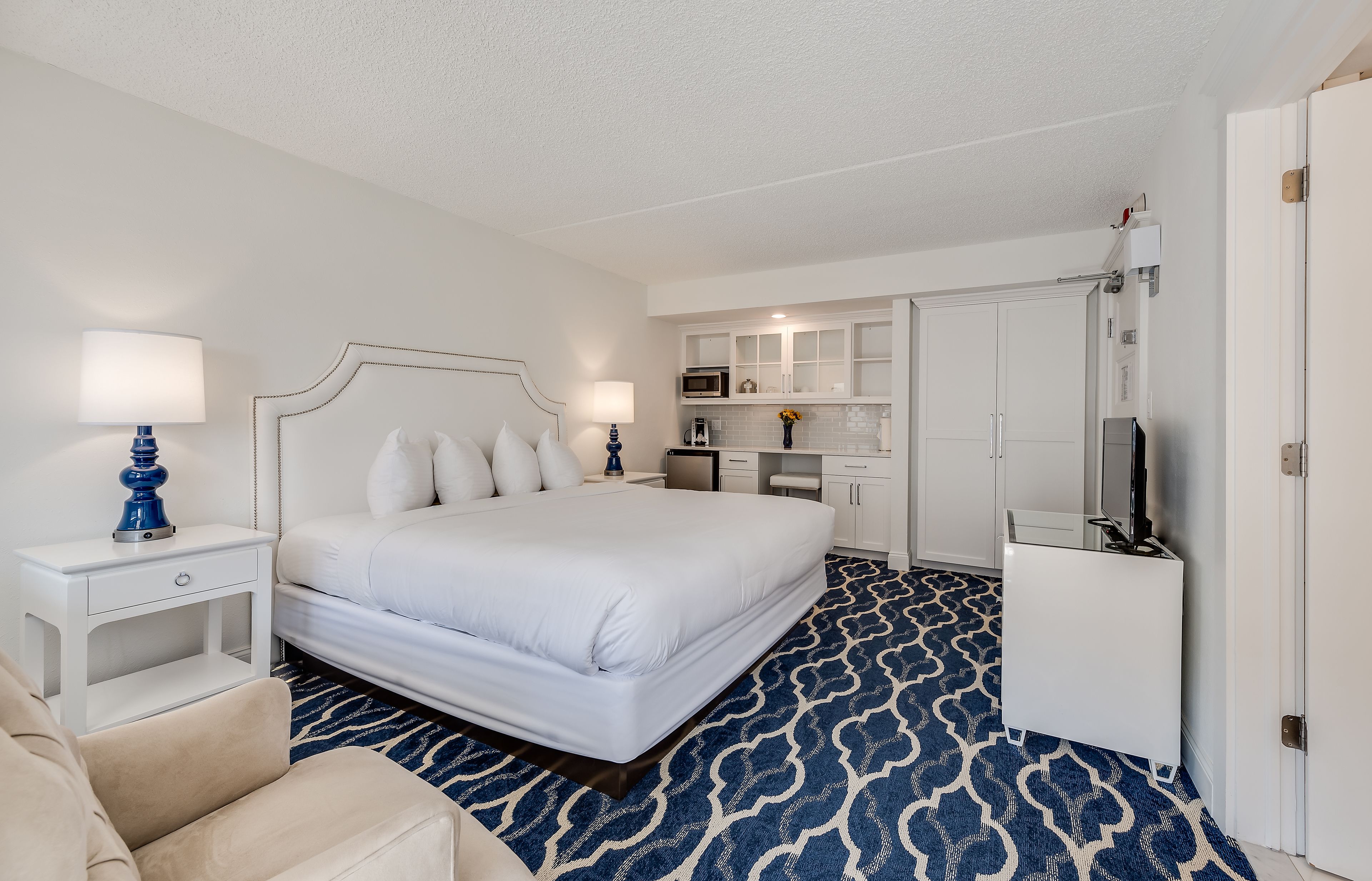 A hotel room with a large bed, a bedside table with a lamp, a small desk, a chair, a kitchenette, and a TV.