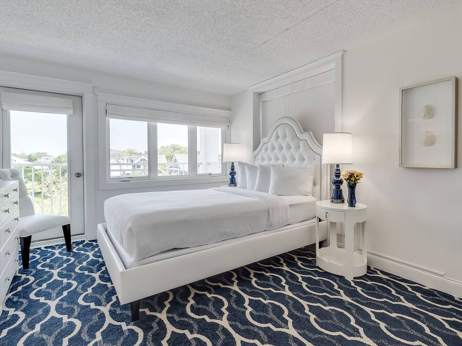 Ein Schlafzimmer mit einem weißen Bett, einem blauen Teppich mit Muster, einer weißen Kommode und einem Fenster mit Blick ins Freie.