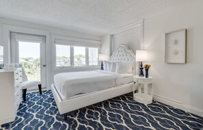 Un dormitorio con una cama blanca, un tapete azul estampado, un tocador blanco y una ventana que da a vistas del exterior.