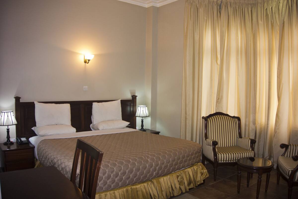 A hotel room with a large bed, two bedside lamps, a striped armchair, a small round table, and a window with curtains.