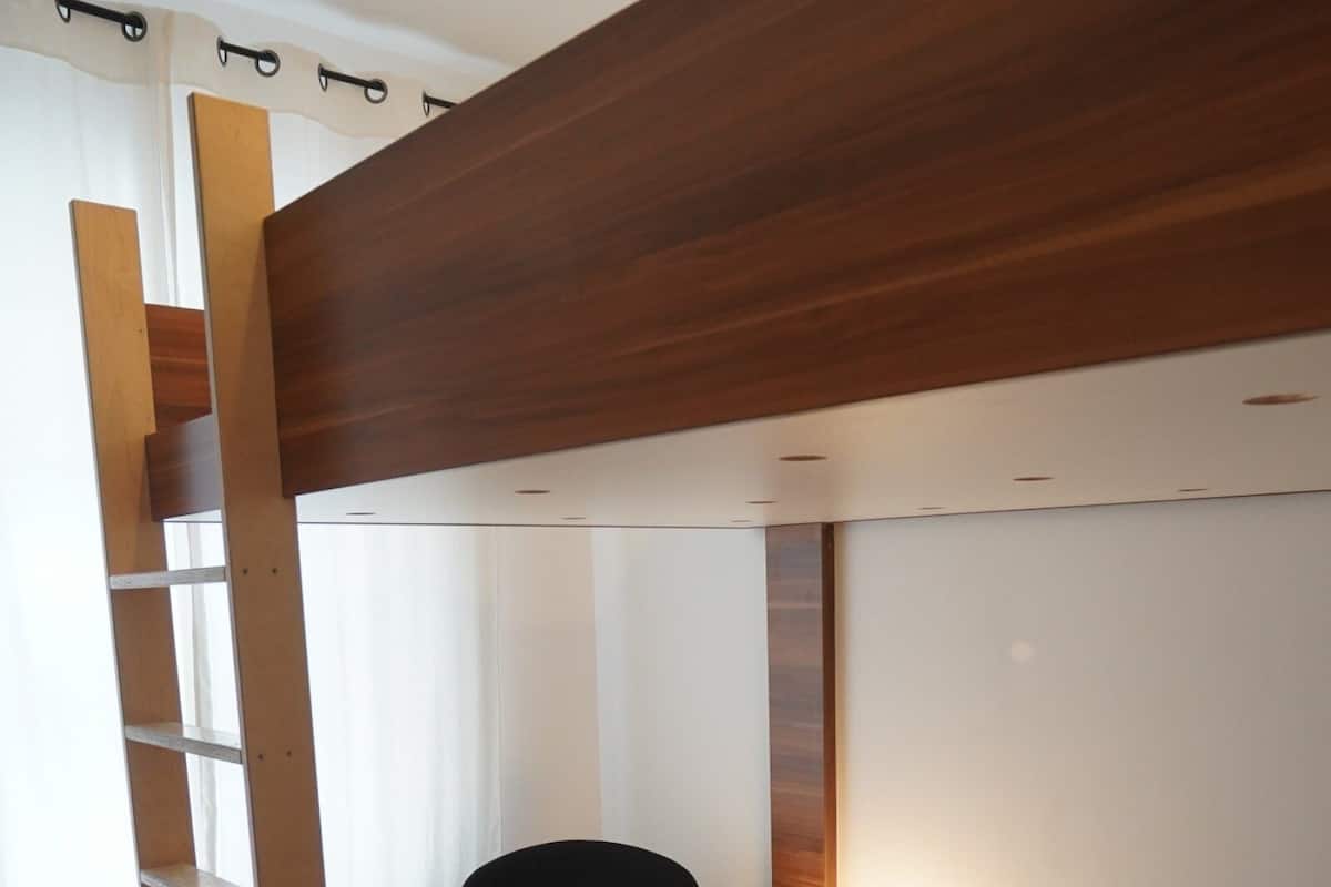 Een kamer met een houten stapelbed, twee zwarte stoelen, een tafeltje met een lamp en een boekenkast.