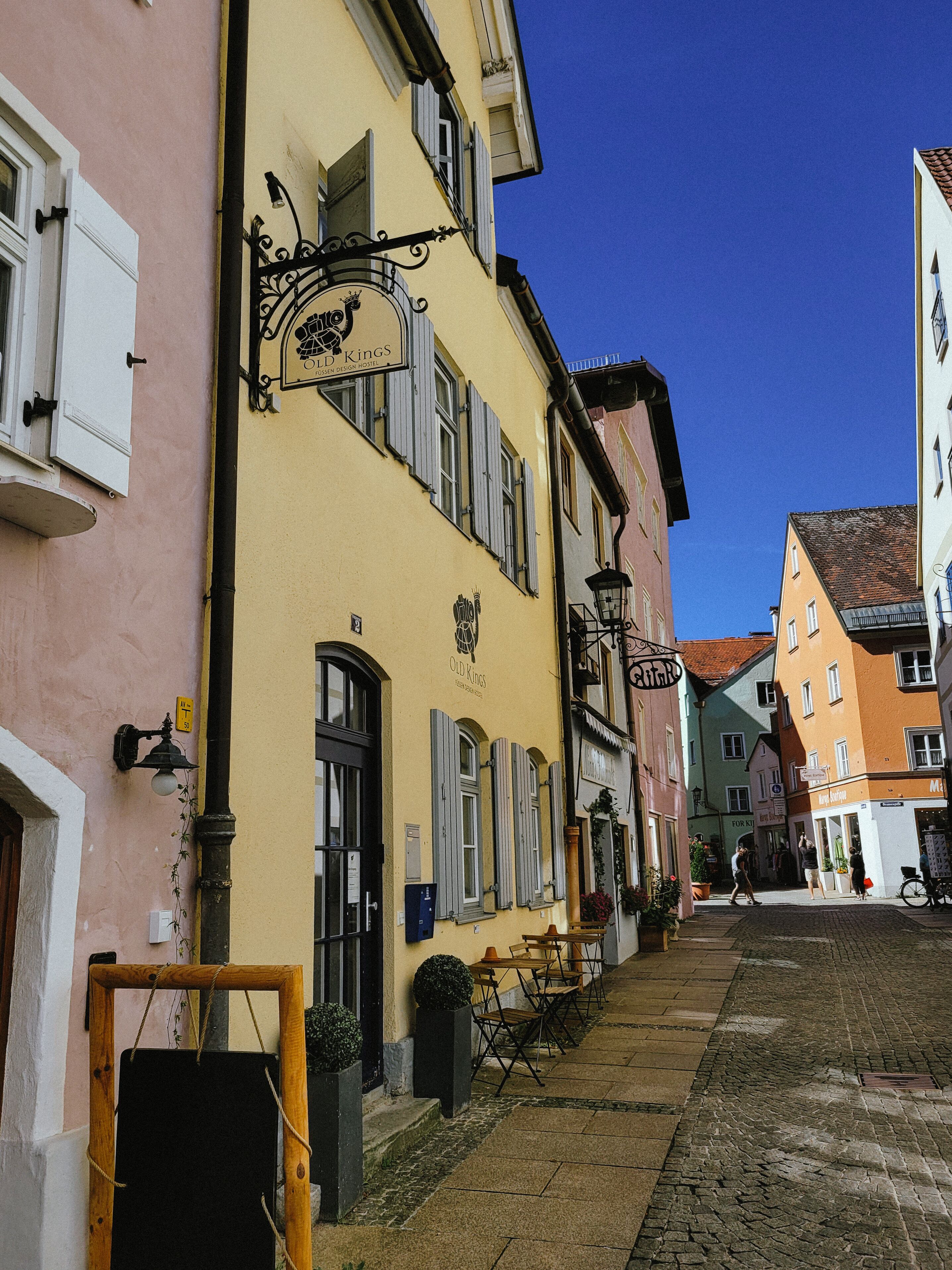Une ruelle étroite pavée, bordée de bâtiments aux couleurs vives, dont un bâtiment jaune portant une enseigne avec l’inscription « Old Kings » et un tableau noir placé à l’extérieur.