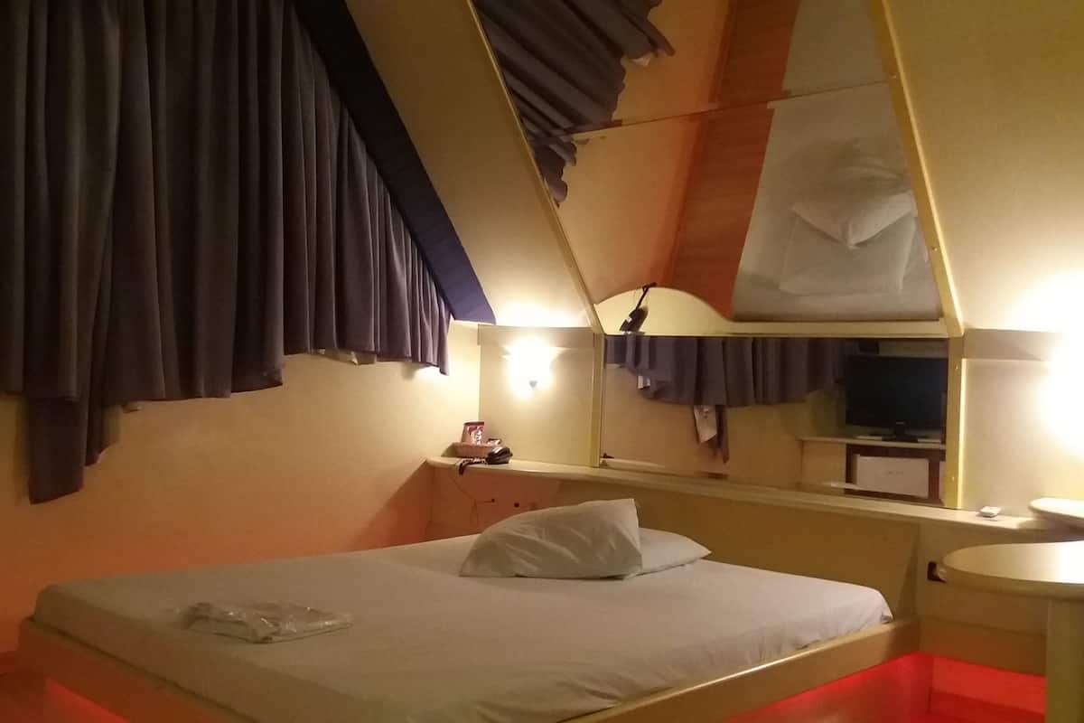 Habitación de hotel con cama, mesita de noche, espejo, televisión y zona de baño.