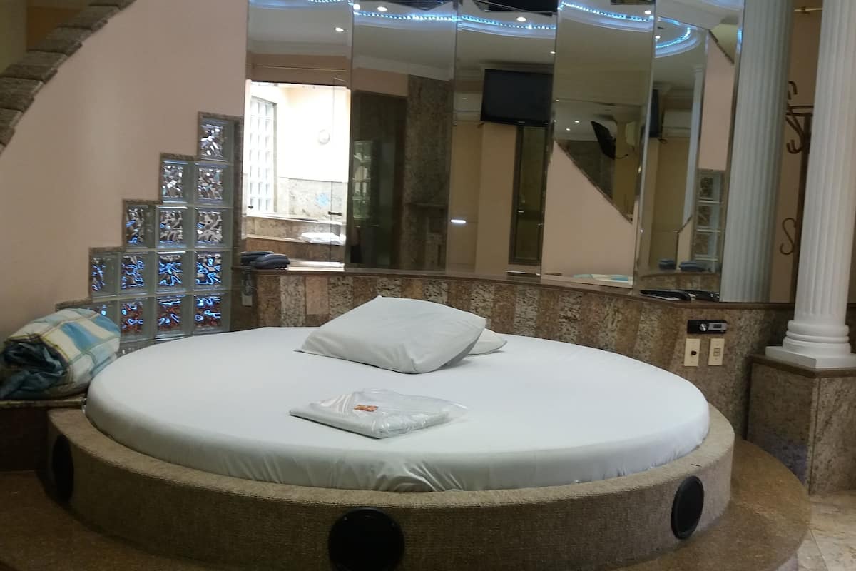 Una cama redonda con colchón y almohadas blancos, un espejo grande y una pared de piedra con una ventana.
