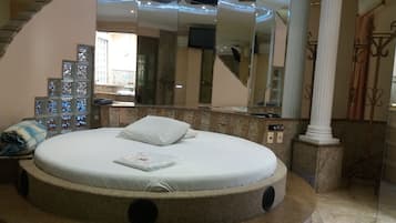 Una cama redonda con colchón y almohadas blancos, un espejo grande y una pared de piedra con una ventana.