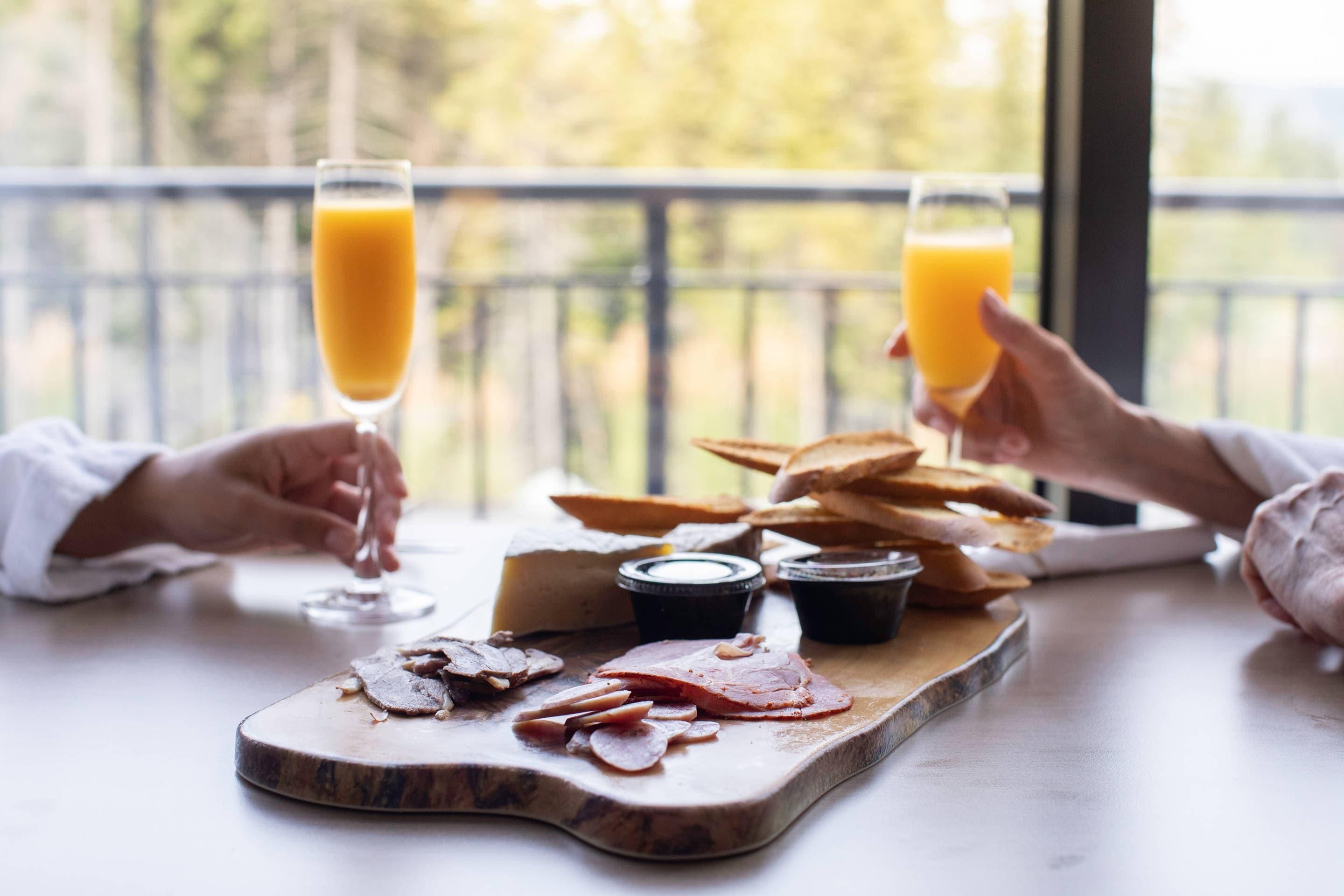 A dining table with a wooden board holding sliced meats and cheeses, two glasses of orange juice, and a plate of bread.
