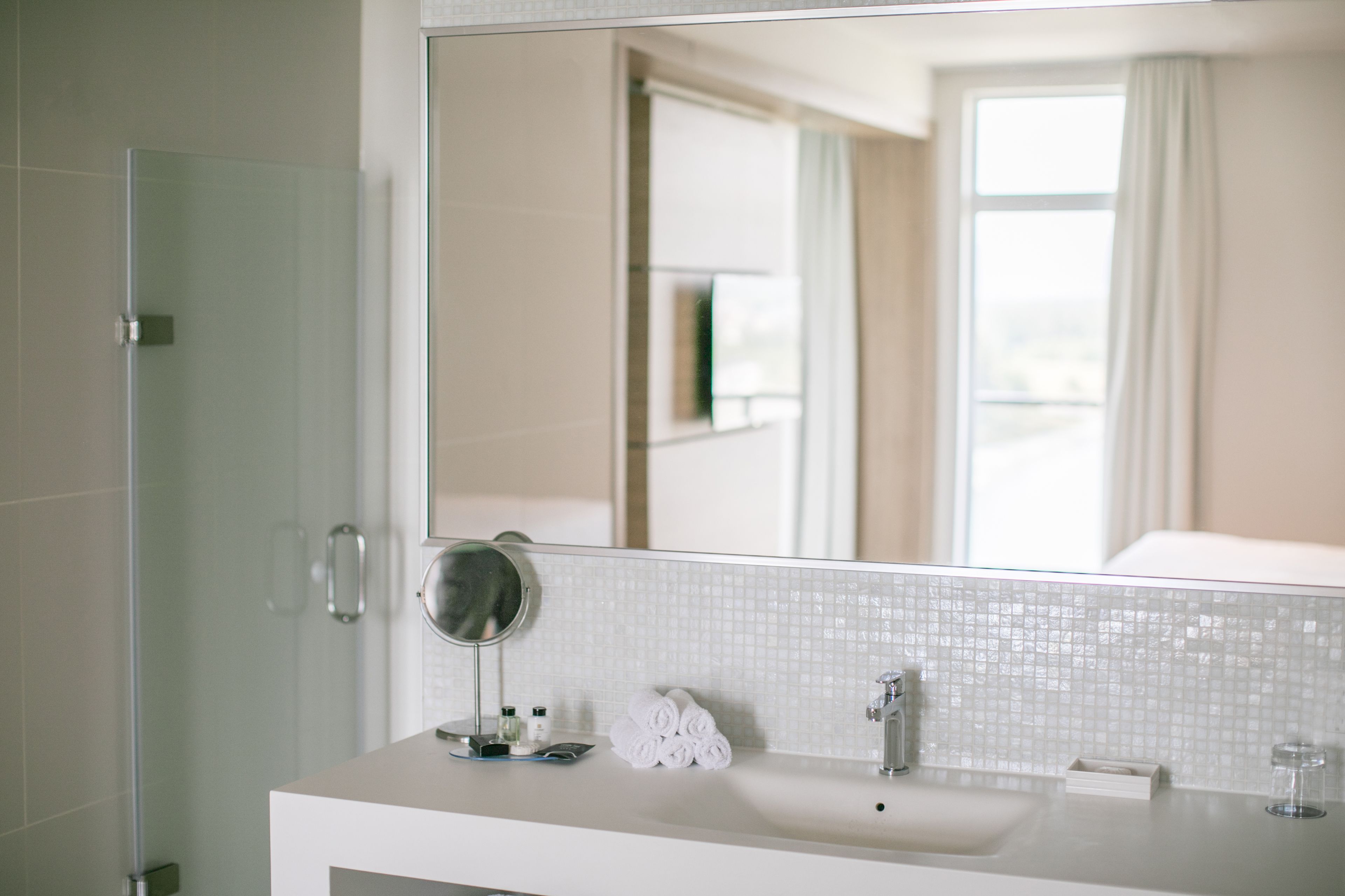 A modern bathroom with a large mirror, white tiles, a sink, and a glass shower enclosure.