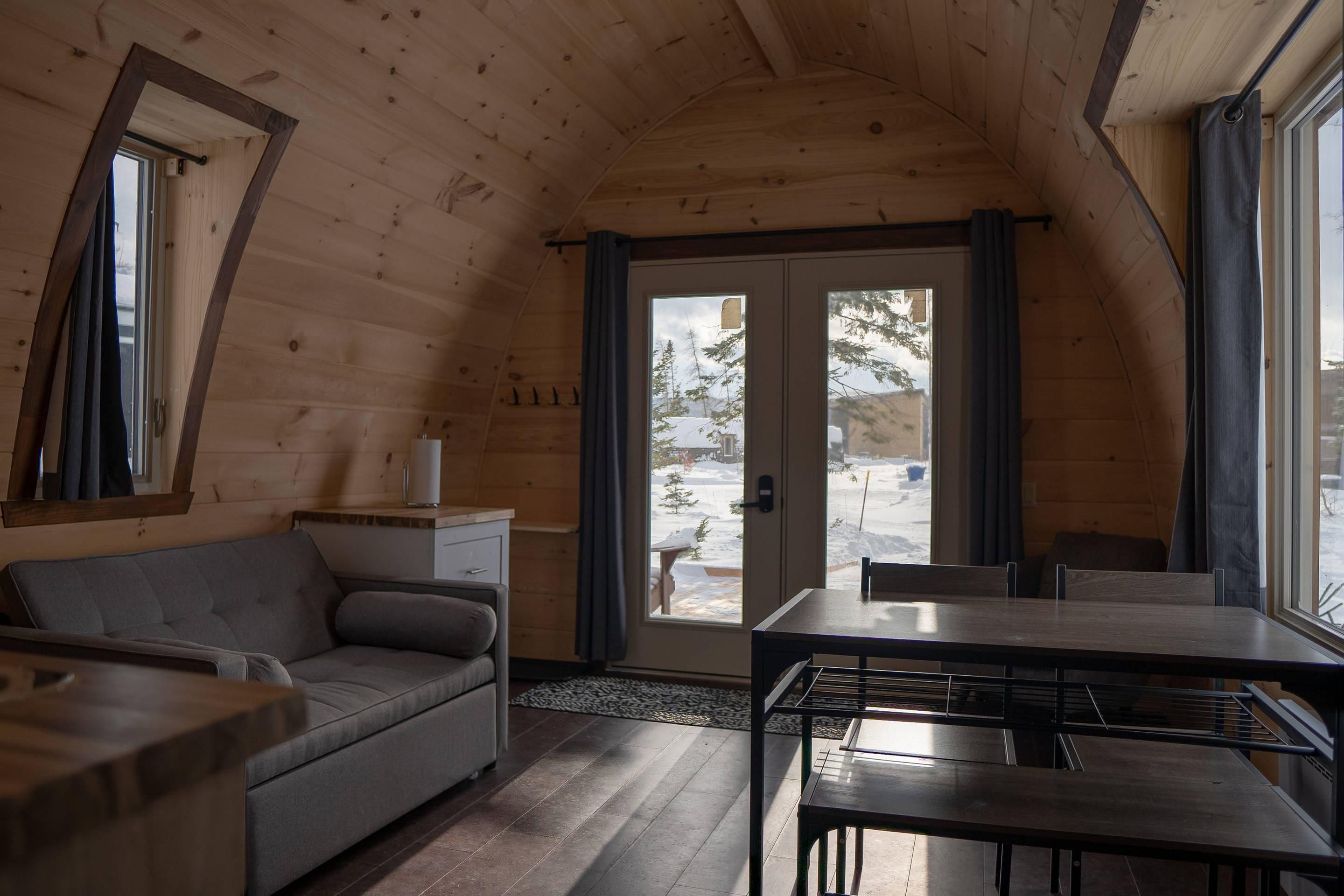 A wooden cabin interior with a sofa, dining table, and a view through the door.