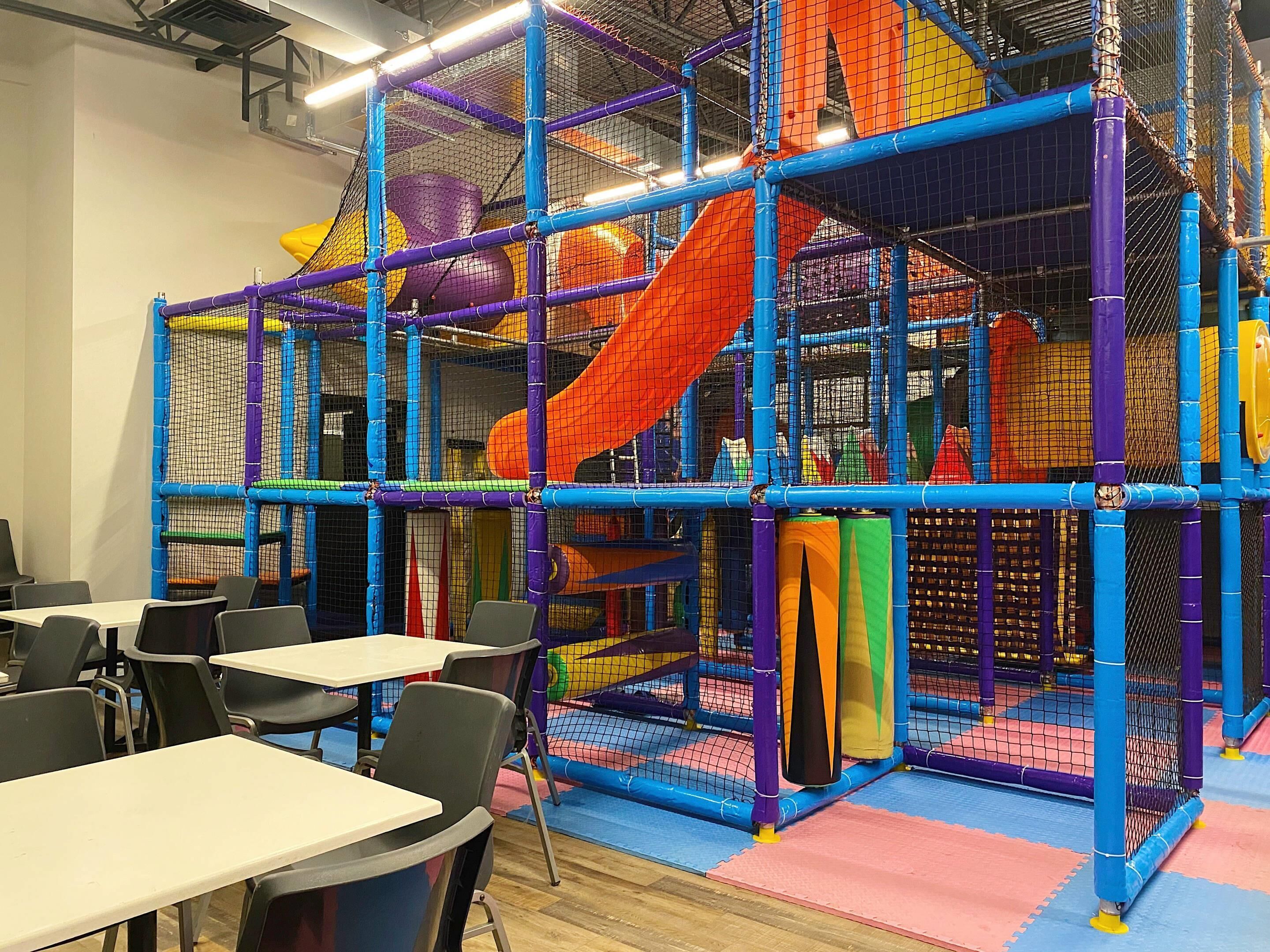 Un espace de jeux avec des toboggans colorés et des structures en filet, entouré de tables et de chaises.