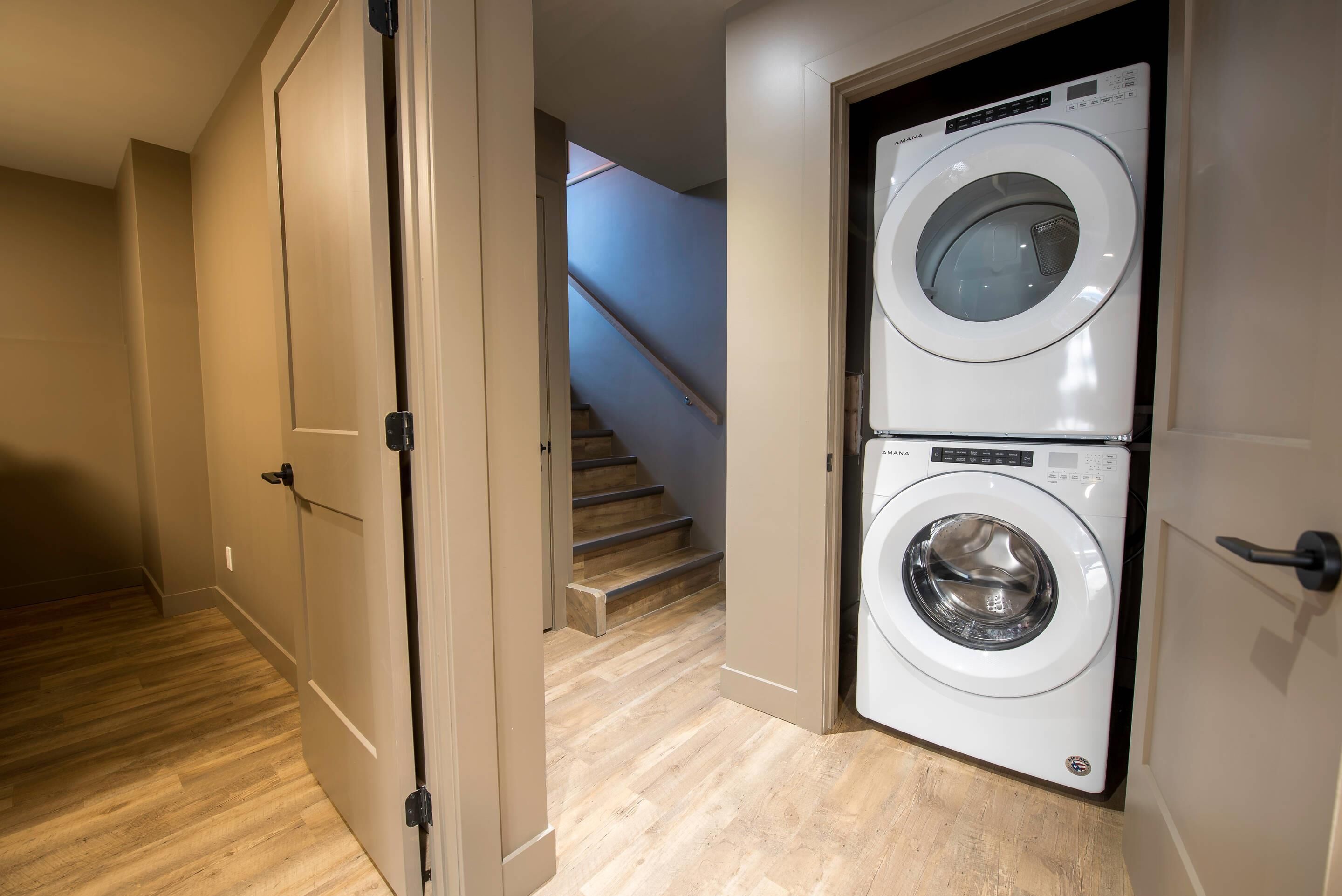 A laundry room with a front-loading washer and dryer, wooden flooring, and a staircase leading to another level.