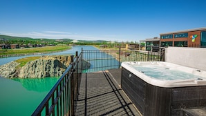 Un bain à remous sur une terrasse donnant sur une rivière, avec un bâtiment moderne en arrière-plan.