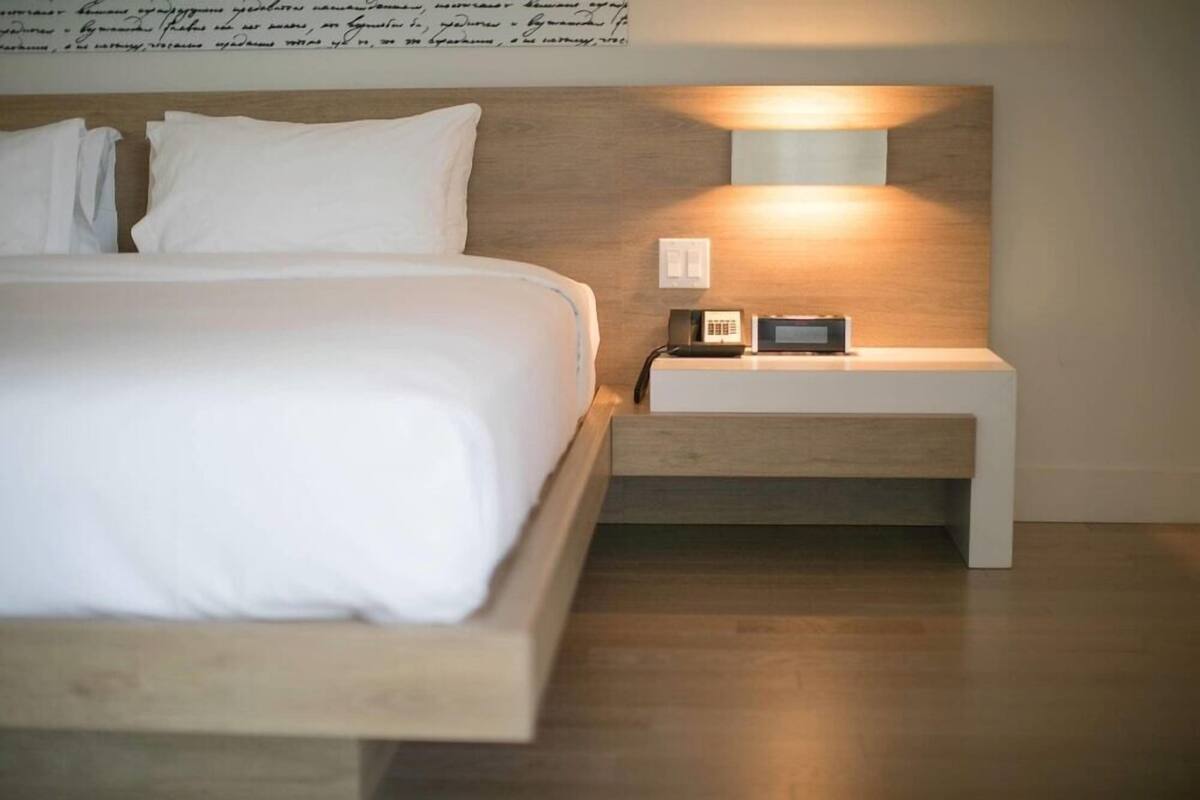 Een netjes opgemaakt bed met witte lakens, een nachtkastje met een lamp en een aan de muur bevestigde telefoon.