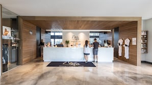 Un espace d’accueil avec un comptoir de réception en bois, un tapis noir orné du nom du spa, et un mur en bois agrémenté d’une bibliothèque et d’un présentoir de peignoirs blancs.
