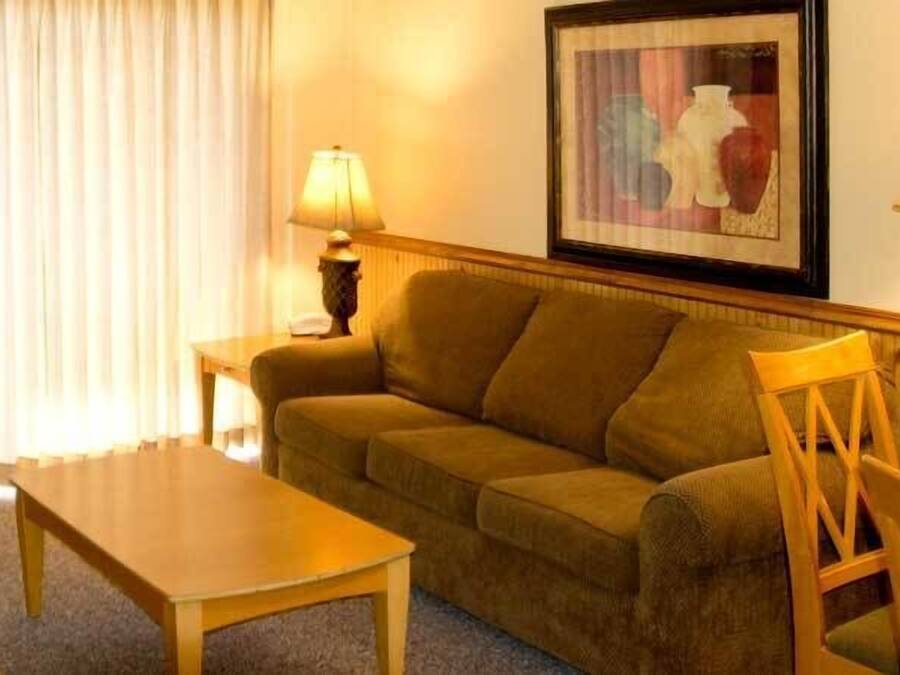 Una habitación con un sofá, una mesa de madera y una silla. Hay una lámpara sobre una mesita auxiliar y un cuadro en la pared.