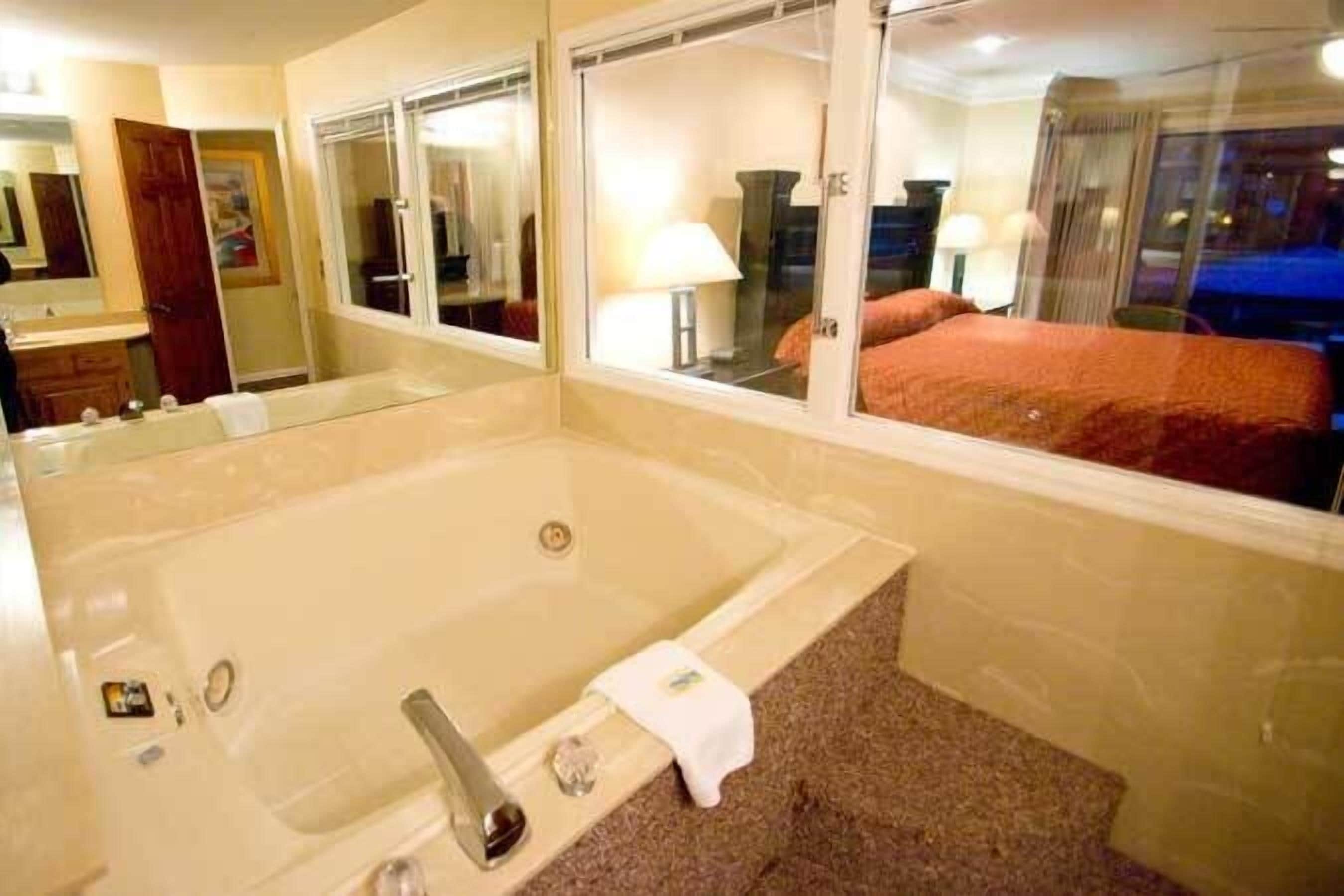A hotel room with a large bathtub, a mirror, and a window with a view of a bed.
