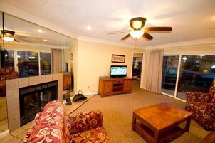 Una sala de estar con chimenea, ventilador de techo, televisor y un amplio ventanal.