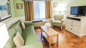 Un salon avec un canapé vert, un fauteuil beige, une table basse en bois, un téléviseur posé sur un meuble blanc et une fenêtre avec des rideaux.