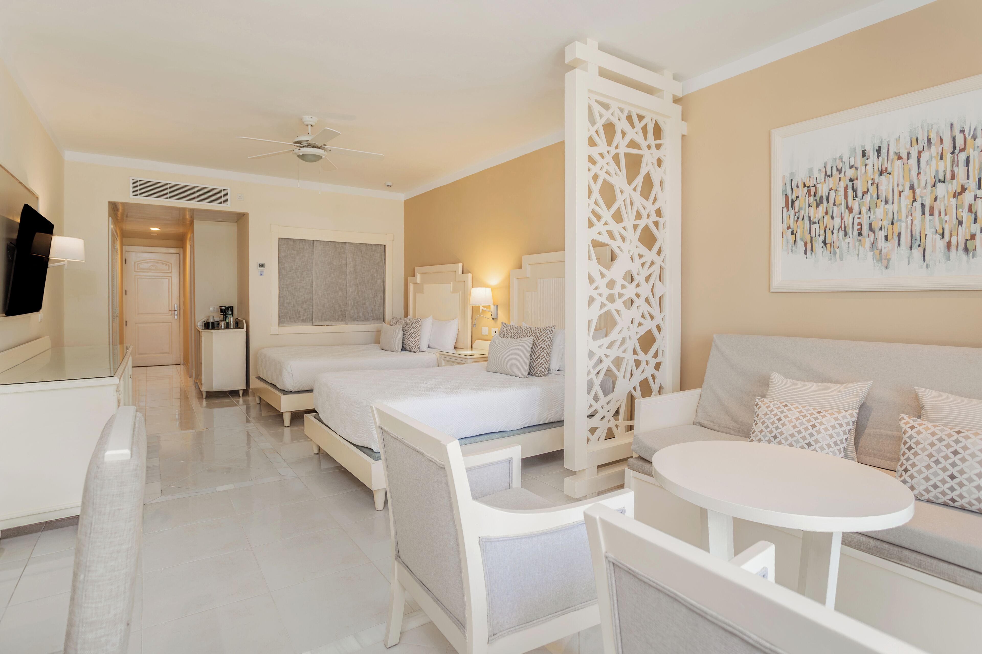Quarto de hotel com duas camas, área de estar com uma mesa, ventoinha no teto, divisória decorativa e um quadro na parede.
