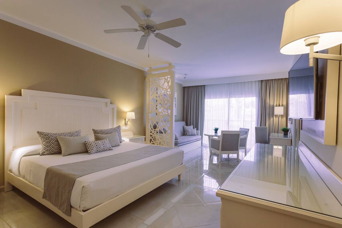 Une chambre d’hôtel avec un grand lit, un bureau, une chaise, un ventilateur de plafond et un téléviseur.