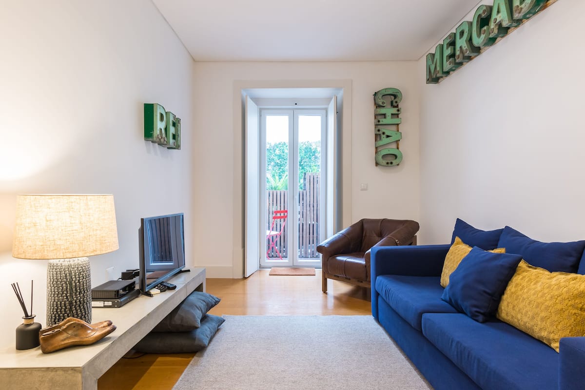 A living room with a blue sofa, a television, a lamp, and a bookshelf. There is a door leading to an outdoor area with a red chair.