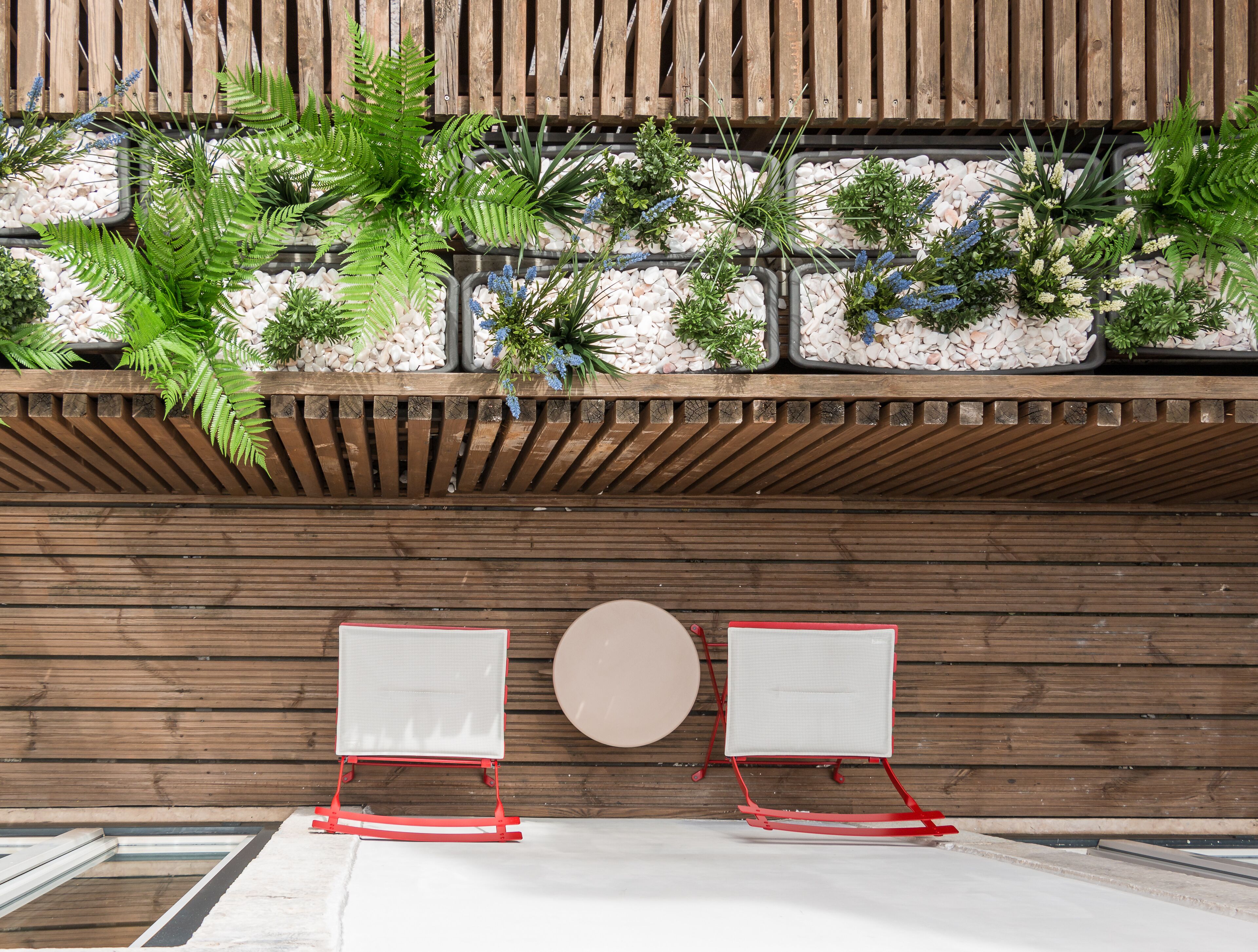 A modern outdoor seating area with two red chairs and a round table, surrounded by a wooden wall and a planter with various plants.