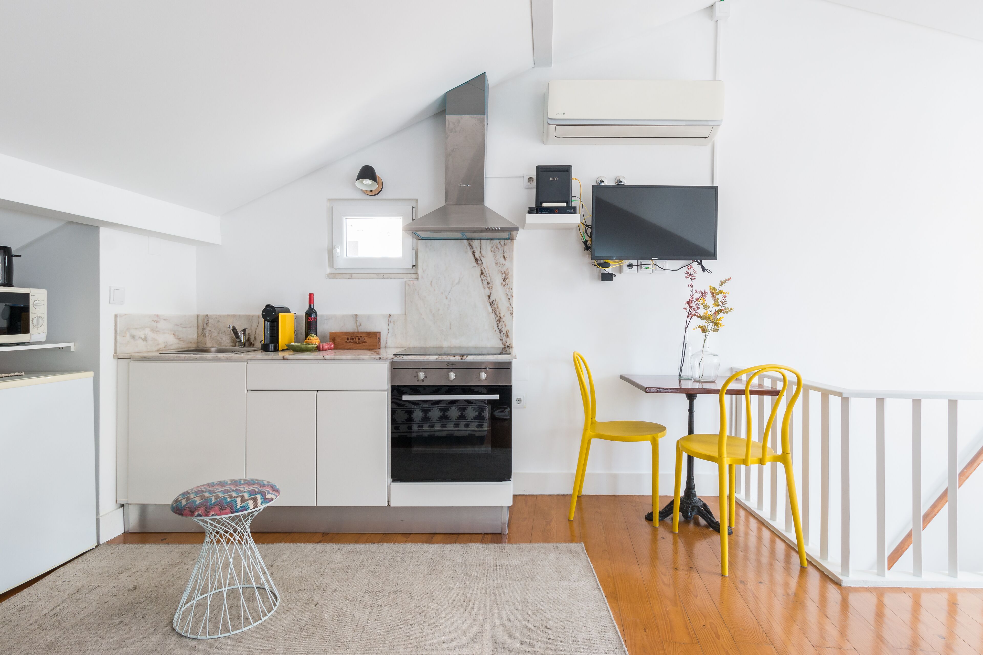A modern kitchen with white cabinetry, a stainless steel range hood, a built-in oven, and a dining area with a small table and two yellow chairs.