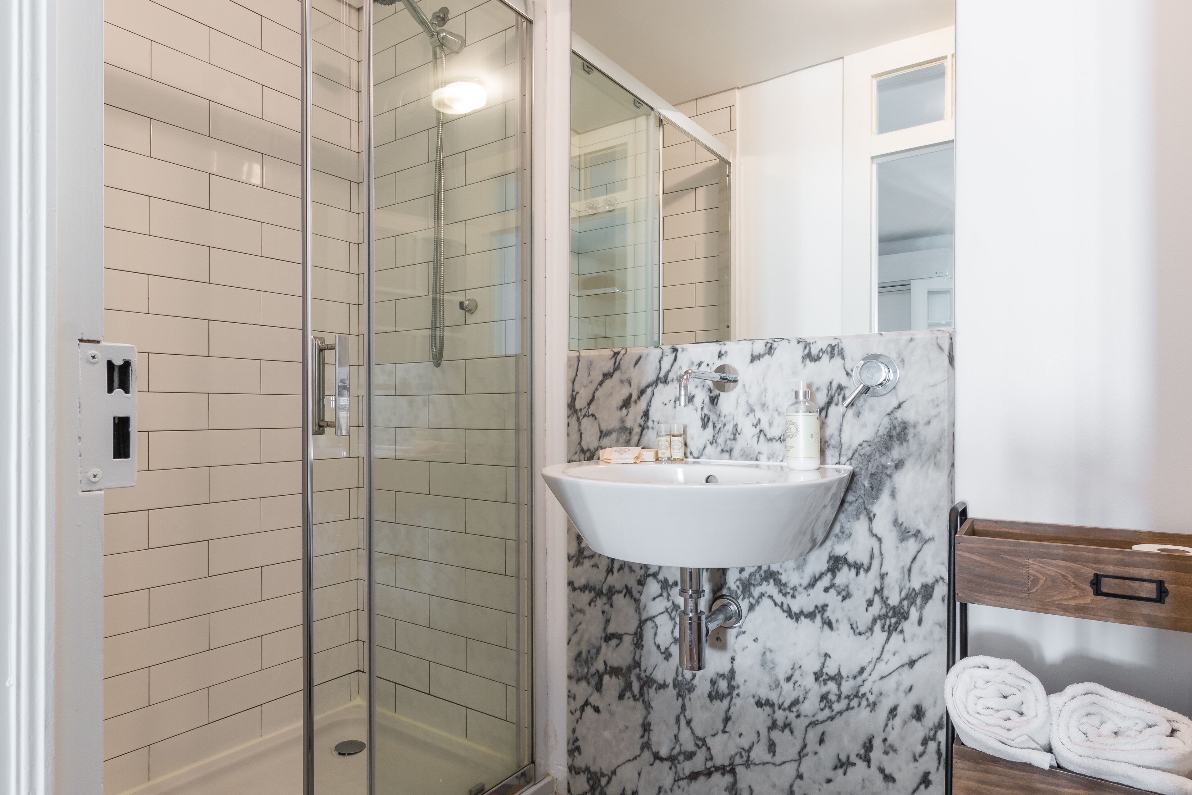 Un baño moderno con ducha de vidrio, un lavamanos blanco y una repisa de mármol, y un perchero de madera.