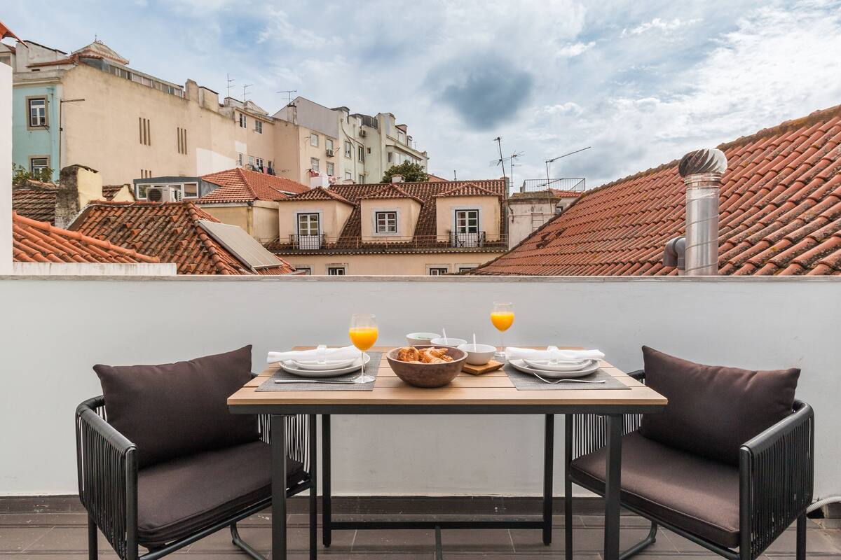 A rooftop terrace with a table set for breakfast, overlooking a cityscape with terracotta rooftops.