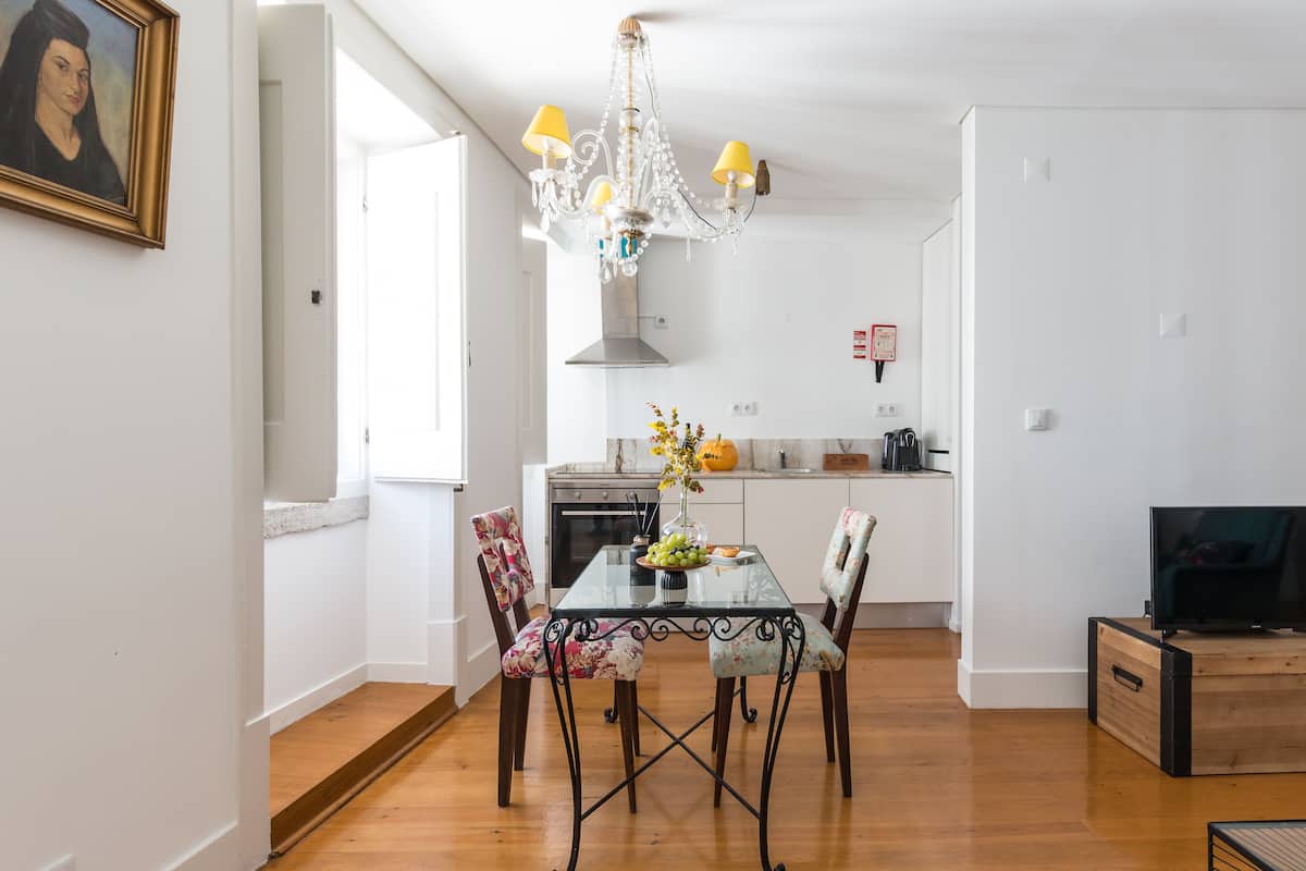 A dining area with a glass table, floral chairs, a chandelier, and a kitchen in the background.