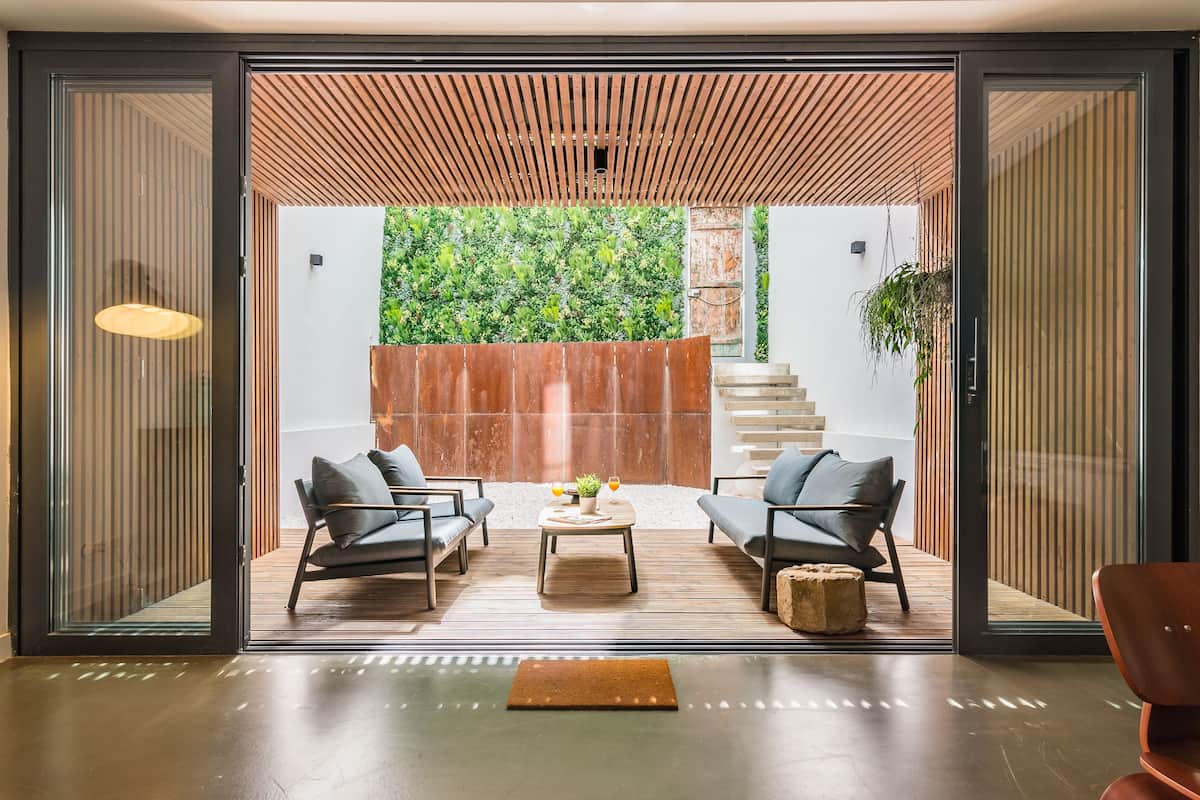 A modern interior with a sliding glass door leading to an outdoor area with a wooden ceiling and a green wall. Inside, there are two black chairs, a wooden coffee table, and a small rug.