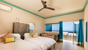 Habitación de hotel con dos camas, ventilador de techo, ventana grande con cortinas y vista al océano.