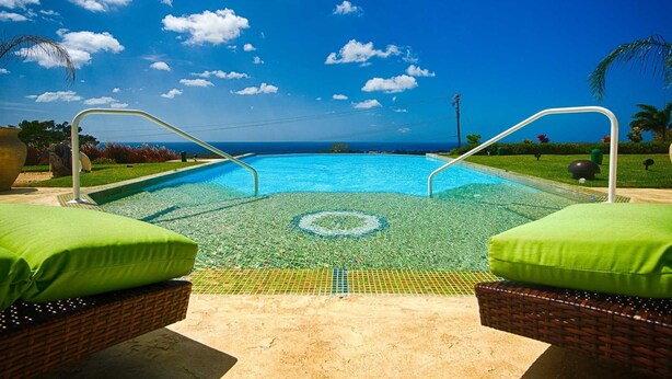 Una piscina con agua azul cristalina, rodeada de cojines verdes y sillas de mimbre, con vistas al océano y al cielo.