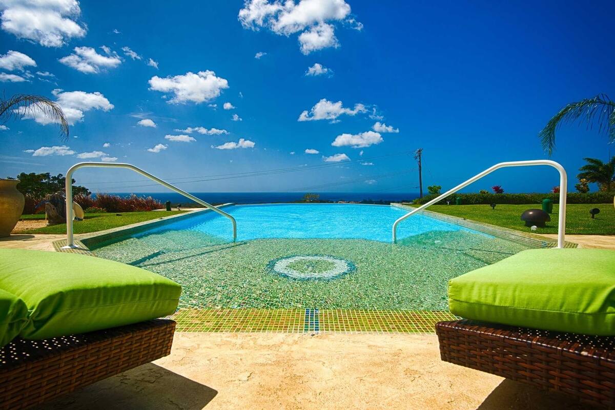 Une piscine à l’eau d’un bleu cristallin, entourée de coussins verts et de fauteuils en osier, offrant une vue sur l’océan et le ciel.