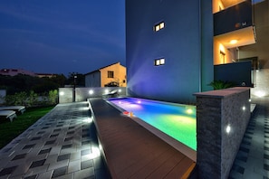 Uma área de piscina ao ar livre moderna, com deck de madeira e paredes de pedra, cercada por um gramado bem cuidado.