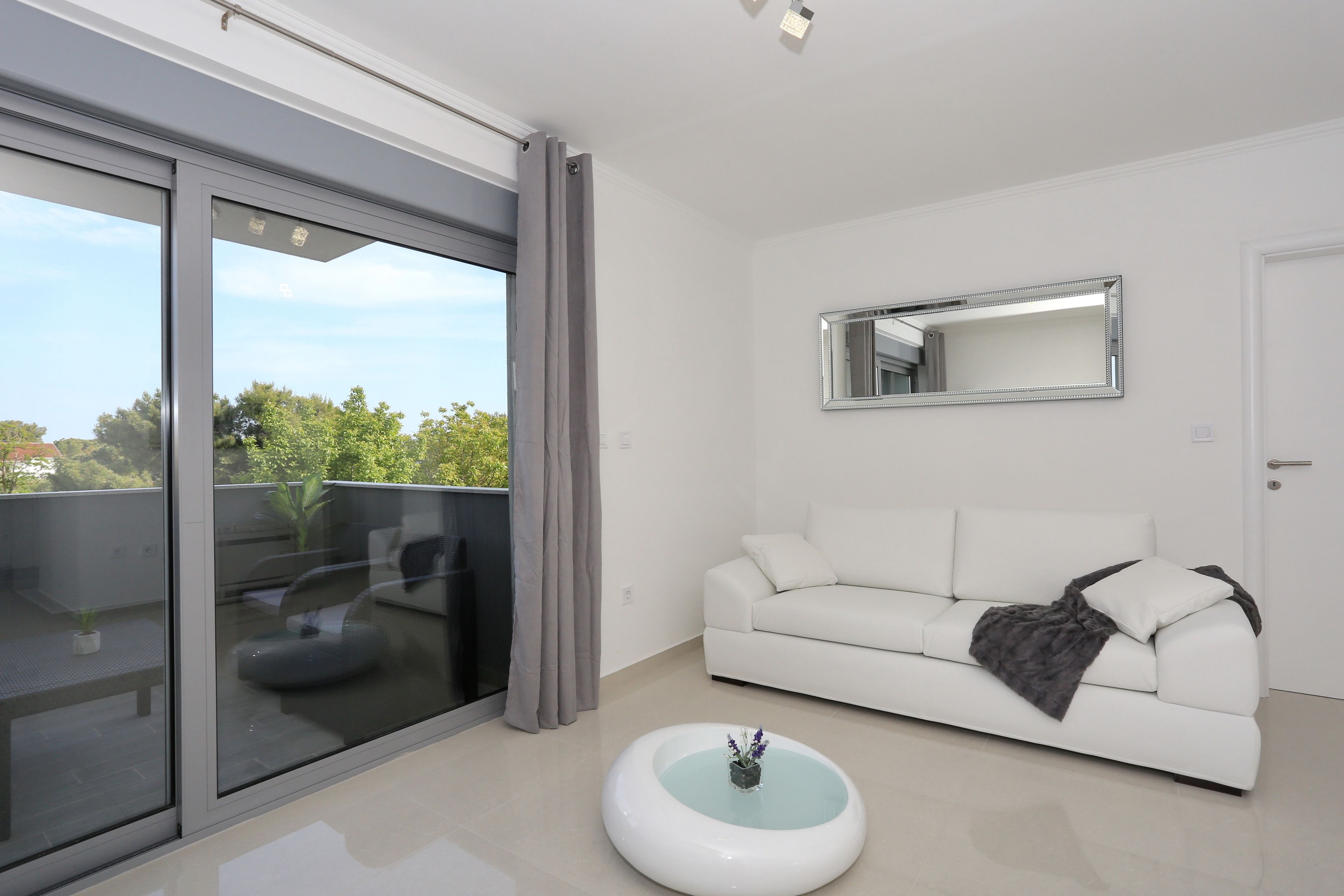 A modern living room with a white sofa, a glass coffee table, and a large sliding glass door leading to an outdoor balcony.
