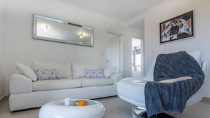 A modern living room with a white sofa, a round coffee table with fruits, a white chair with a blanket, a large mirror, and a painting on the wall. - Sun Resort Nikolina (Bibinje)