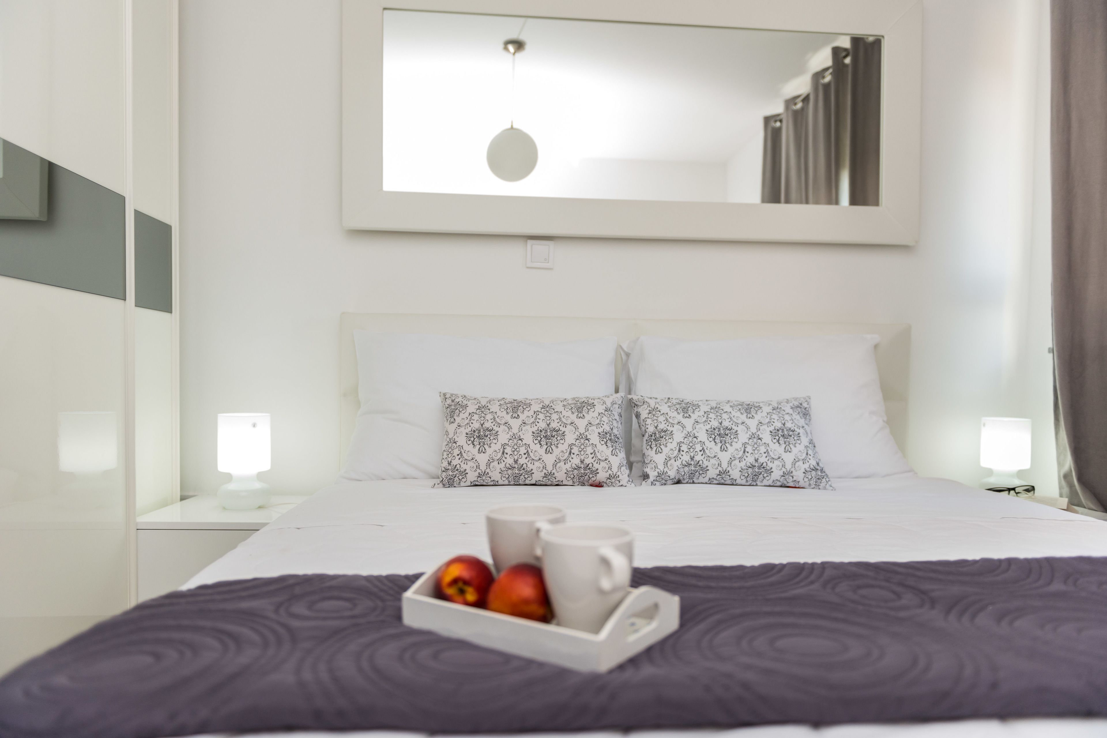 Habitación de hotel con una cama bien hecha, una bandeja con manzanas y una taza, y un espejo grande sobre la cama.
