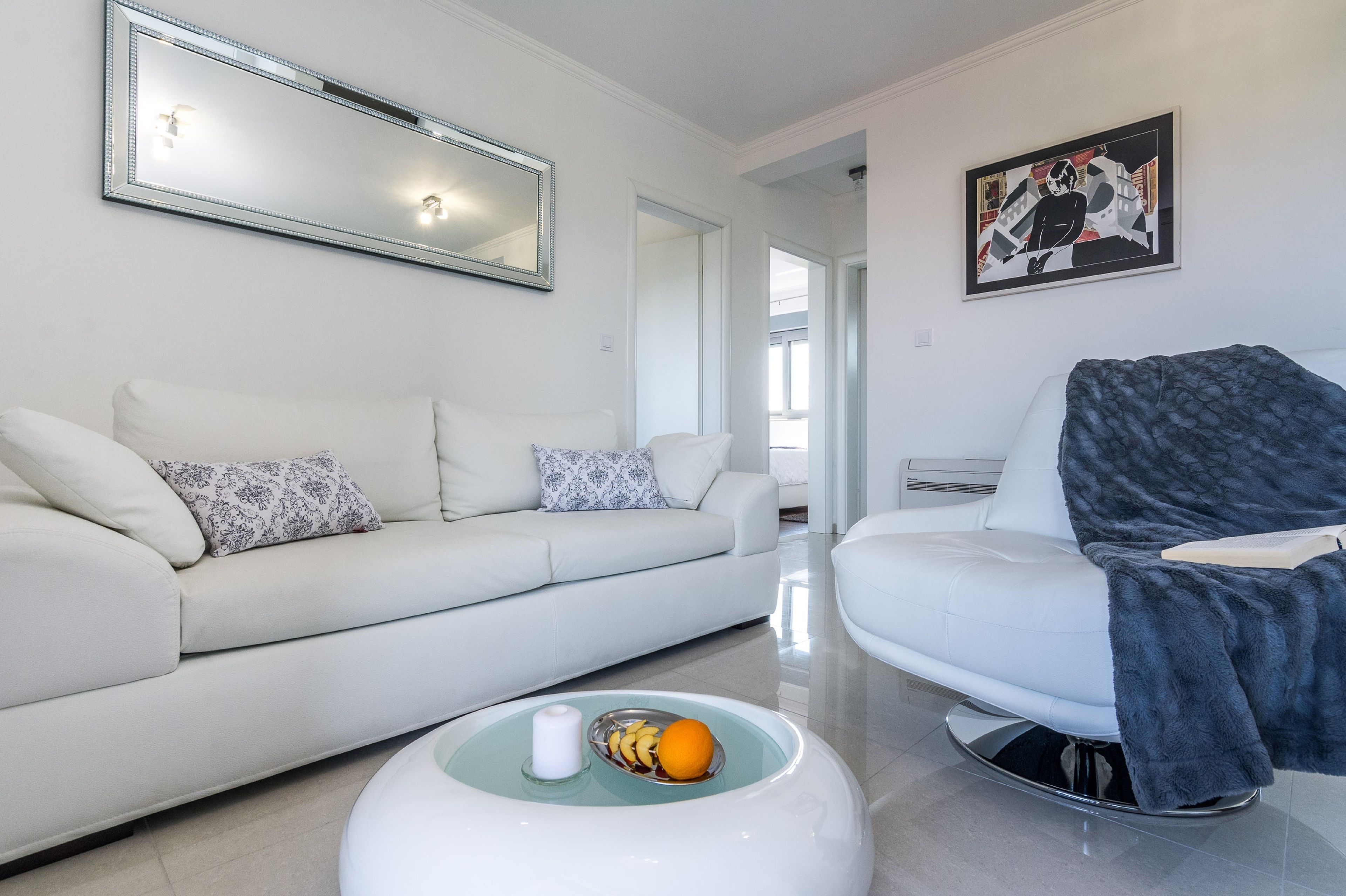 A modern living room with a white sofa, a glass coffee table with fruits and a candle, and a large mirror on the wall.