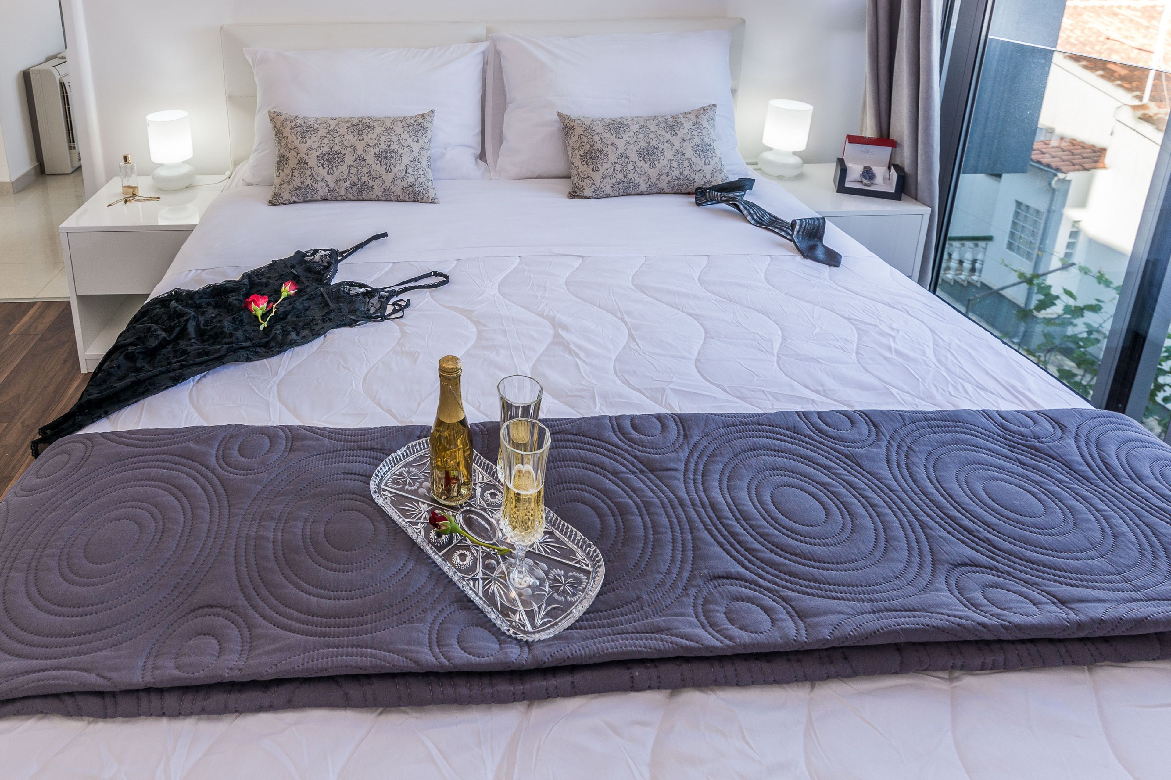 A neatly made bed with a decorative pillow, a bedside table with a lamp, and a tray with a bottle and glass of champagne.