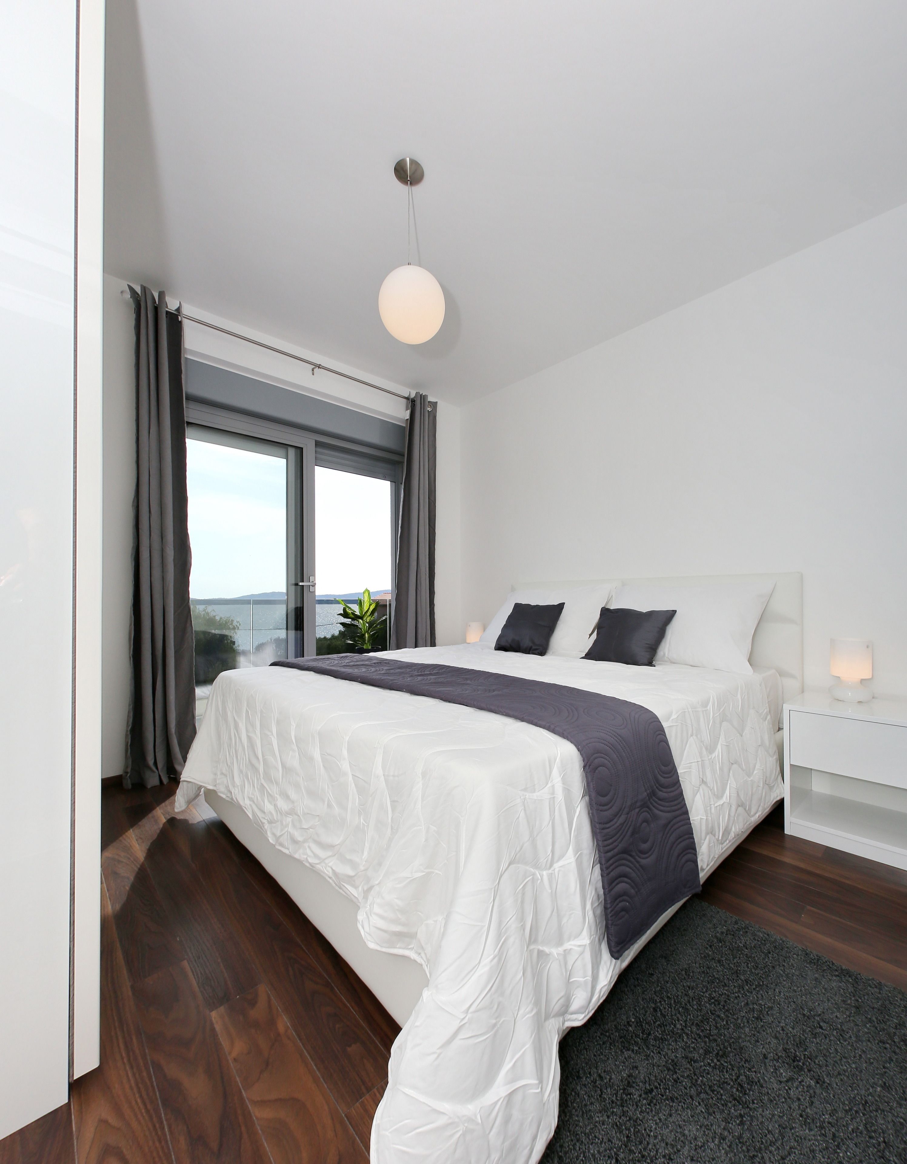 A modern bedroom with a large bed, a bedside table with a lamp, a view of the outdoors through a window, and a hanging light fixture.