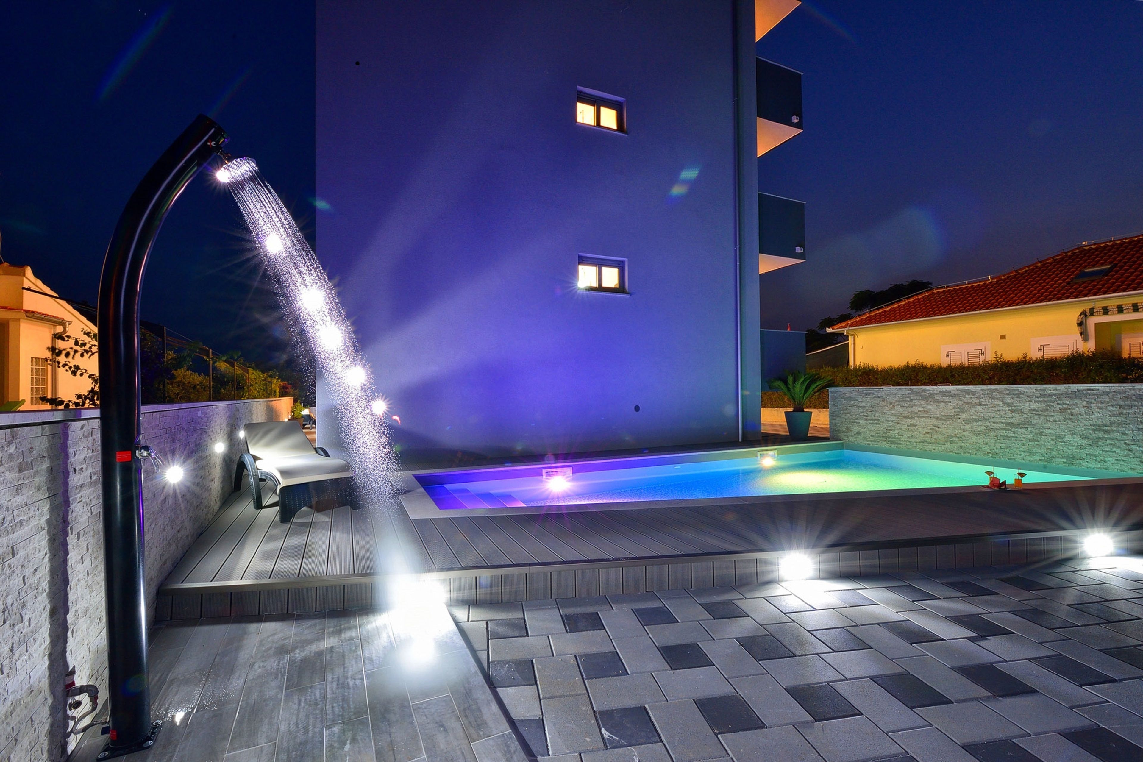 A modern outdoor pool area with a water feature, a lounge chair, and a well-lit building in the background.