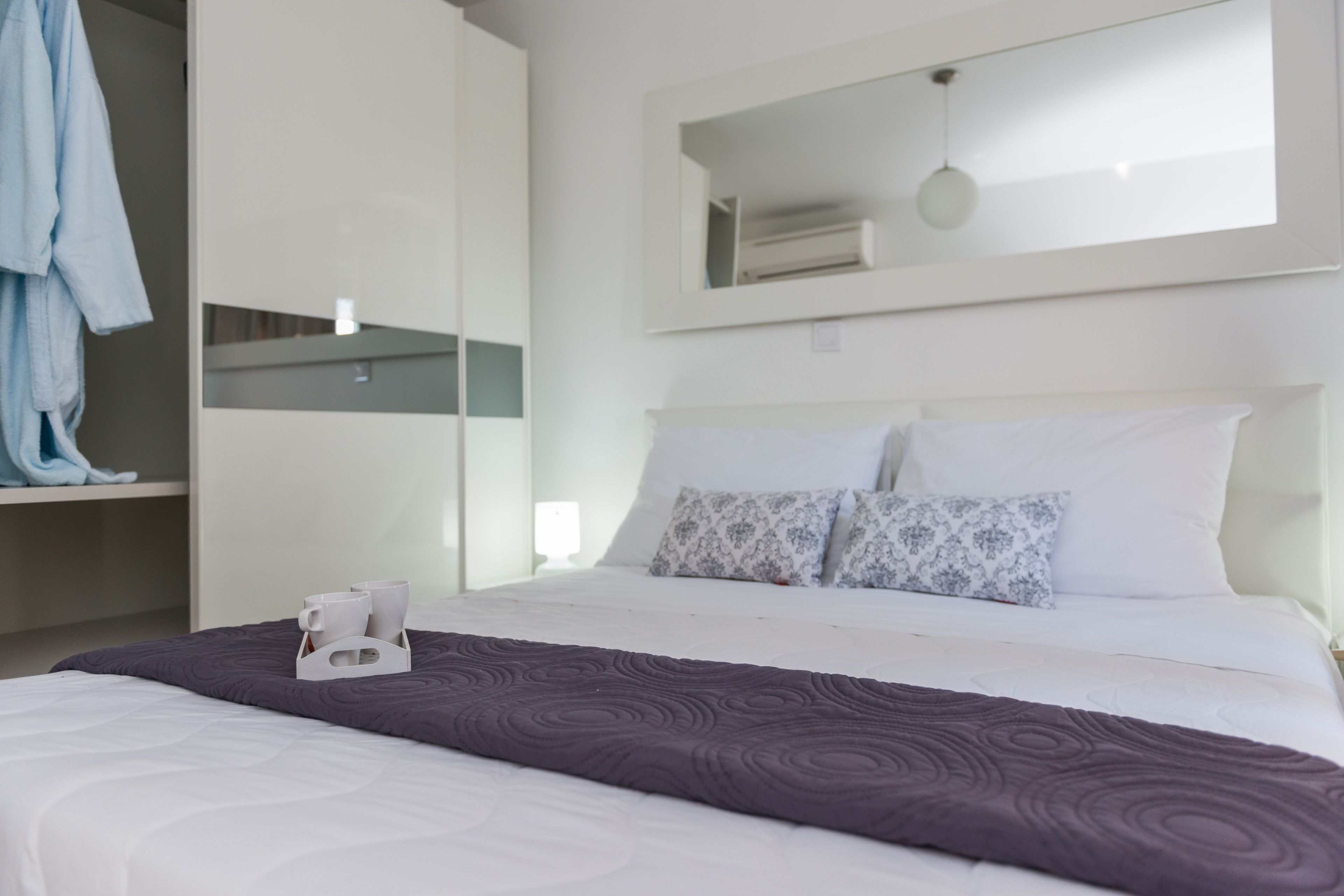 A hotel room with a neatly made bed, a bedside table with a cup holder, a hanging robe, and a large window with a view of the sky.
